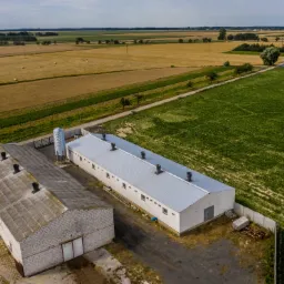 Widok z lotu ptaka na dwa budynki gospodarcze z silosami na tle p&oacute;l uprawnych i łąk w słoneczny dzień. Jeden budynek z szarej cegły, drugi biały z szarym dachem.
