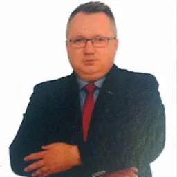 Portret mężczyzny w okularach, granatowym garniturze i czerwonym krawacie, z rękami skrzyżowanymi na piersiach, na jasnym tle.