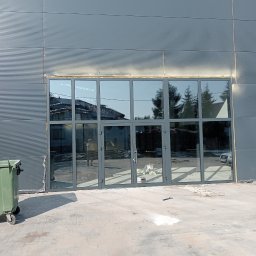 ZUH STOLKON - Szklana fasada budynku przemysłowego z szarymi ramami, widoczne odbicie sylwetki osoby w szybach, obok kontener na śmieci, ślady prac budowlanych na chodniku.