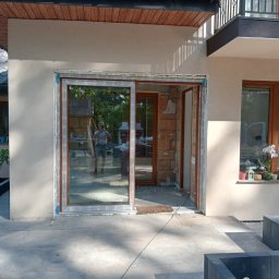 ZUH STOLKON - Montaż przesuwnych drzwi tarasowych z brązową ramą w nowym domu, widoczne cegły i pianka montażowa, odbicie osoby robiącej zdjęcie w szybie.