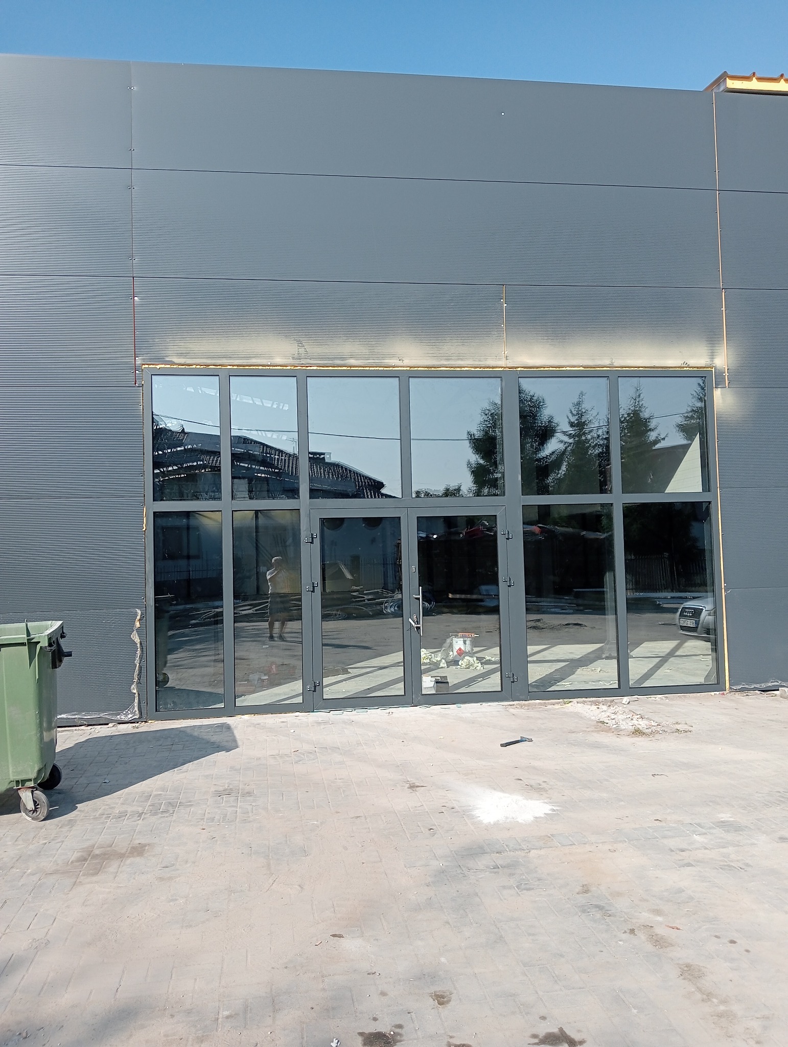 Szklana fasada budynku przemysłowego z szarymi ramami, widoczne odbicie sylwetki osoby w szybach, obok kontener na śmieci, ślady prac budowlanych na chodniku.