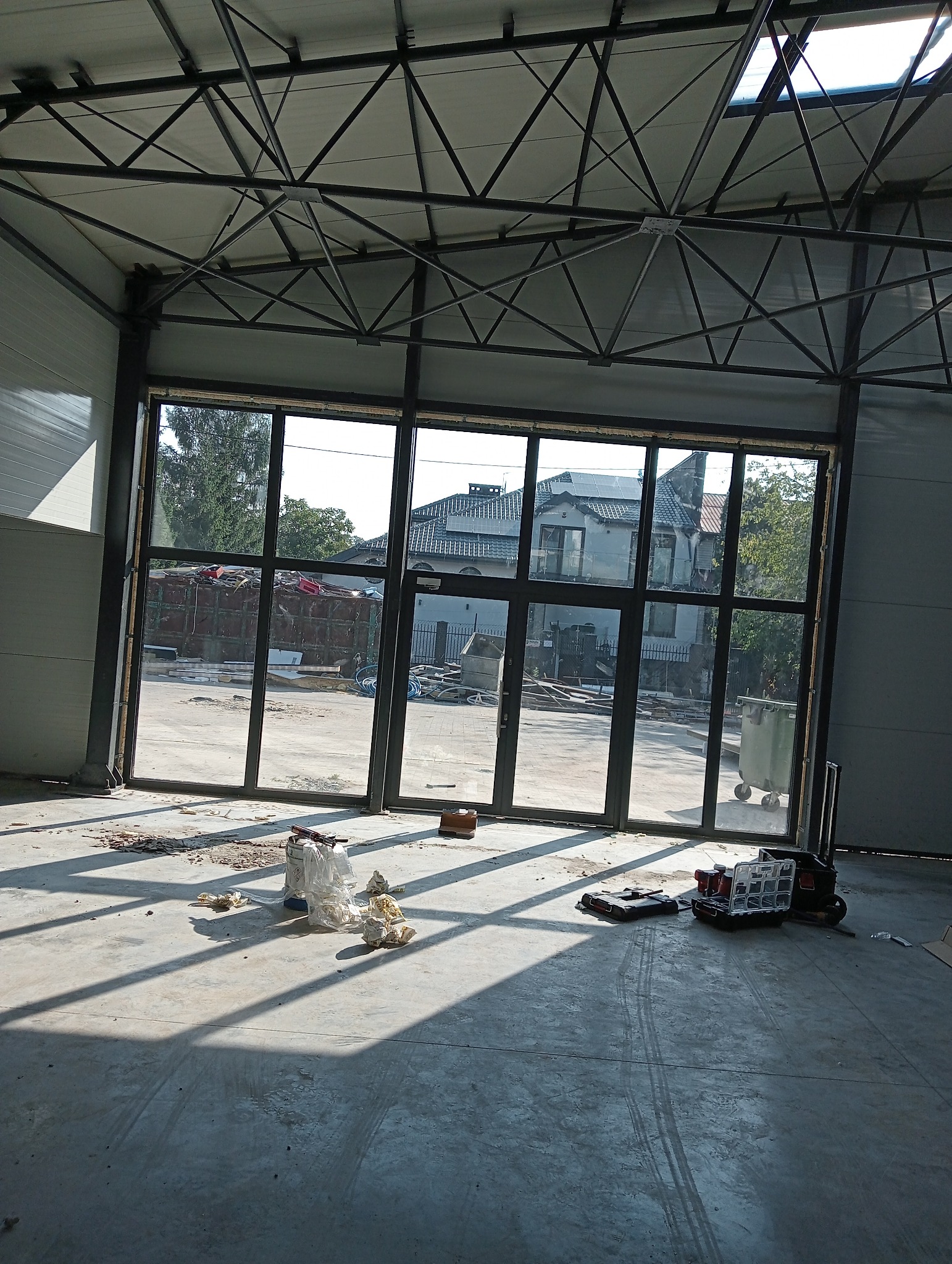 Wnętrze hali produkcyjnej w trakcie montażu dużej, przeszklonej konstrukcji z czarnymi profilami, widoczne narzędzia i materiały budowlane, w tle budynki mieszkalne.