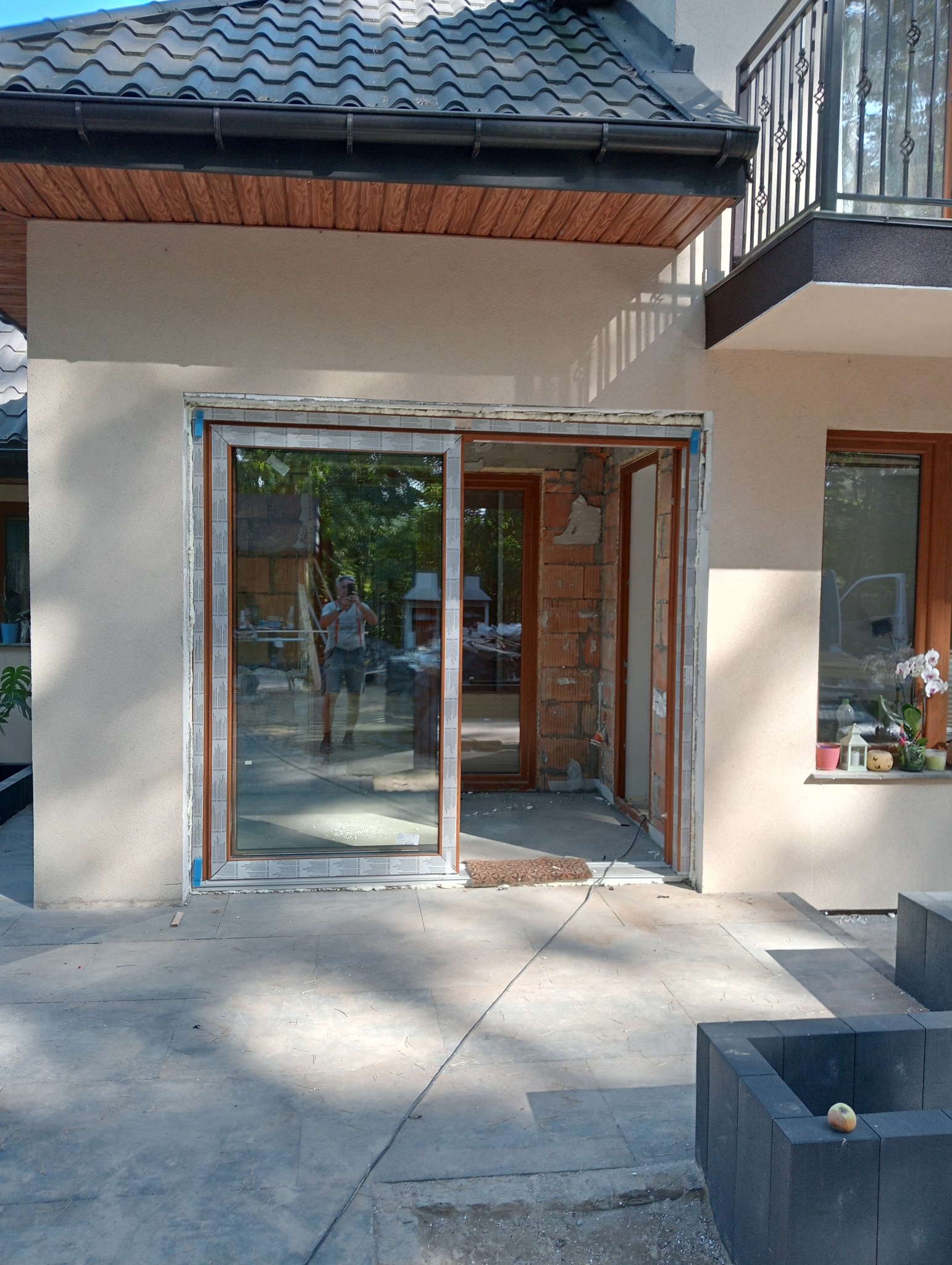 Montaż przesuwnych drzwi tarasowych z brązową ramą w nowym domu, widoczne cegły i pianka montażowa, odbicie osoby robiącej zdjęcie w szybie.