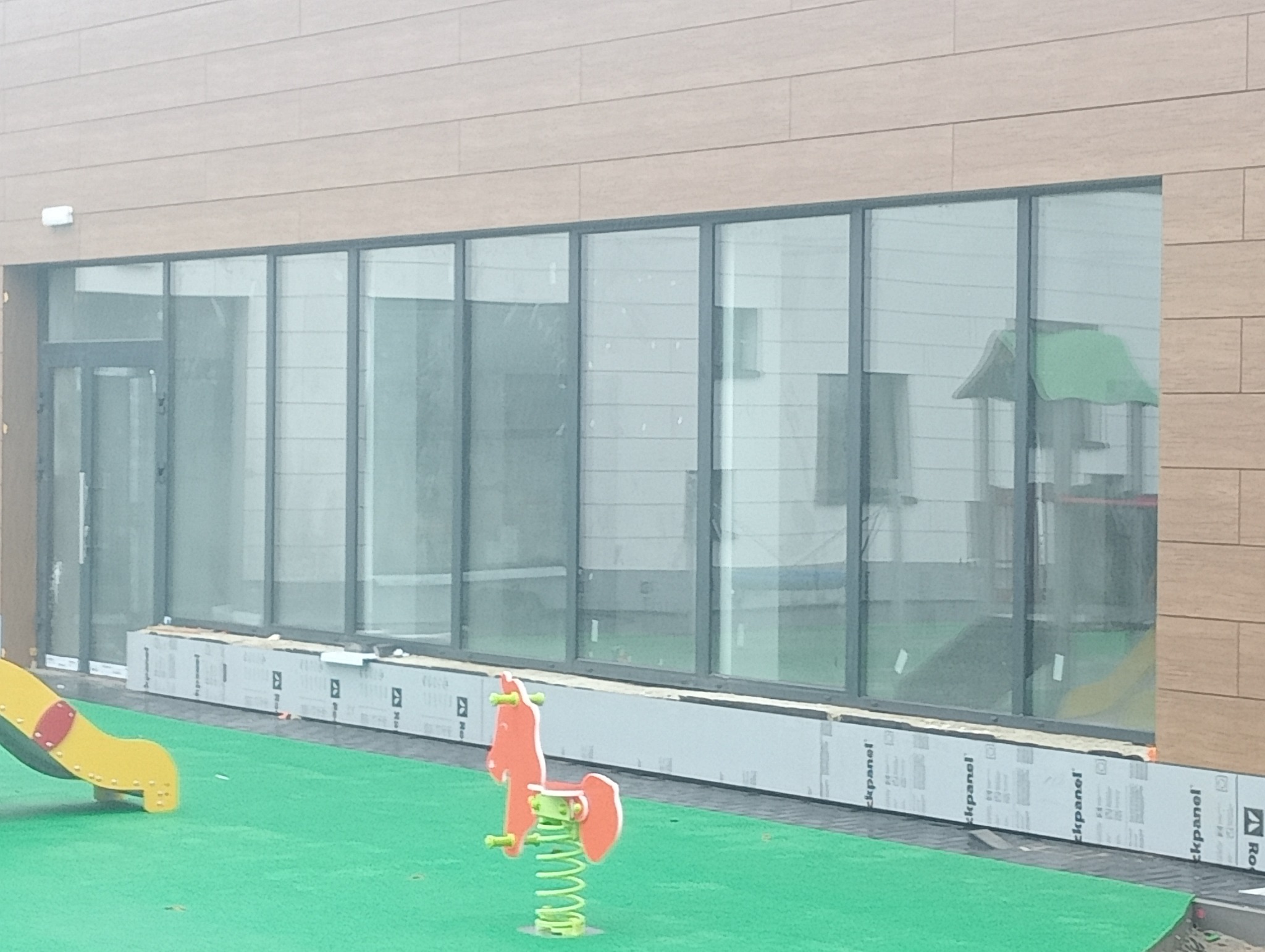 Duże przeszklenia w budynku przedszkola z placem zabaw, widoczne zabawki na sprężynach i zjeżdżalnia, ściany wyłożone panelami imitującymi drewno.