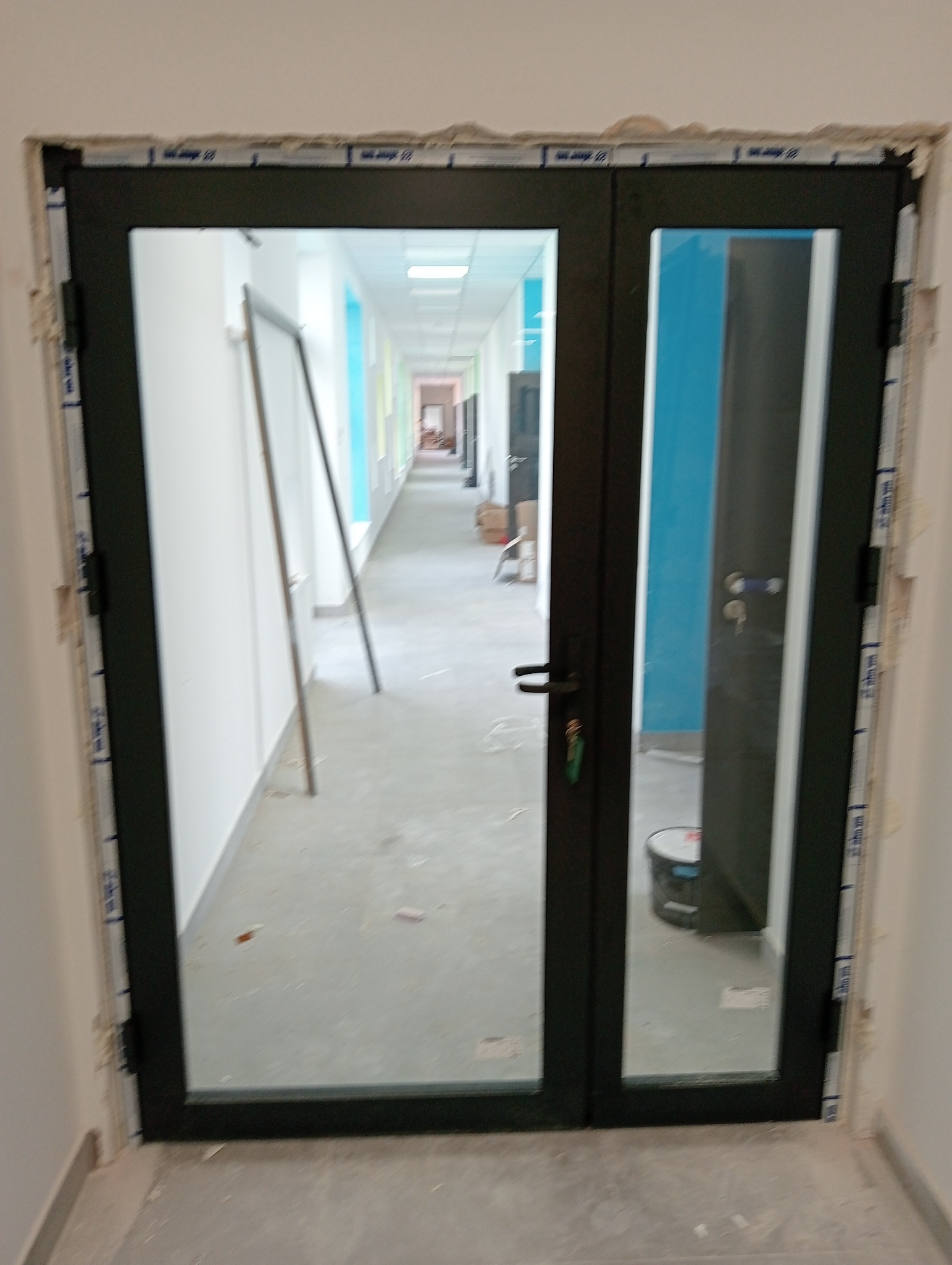 Czarne, dwuskrzydłowe drzwi z widokiem na długi korytarz z kolorowymi ścianami, widoczne ślady montażu na ościeżnicy.