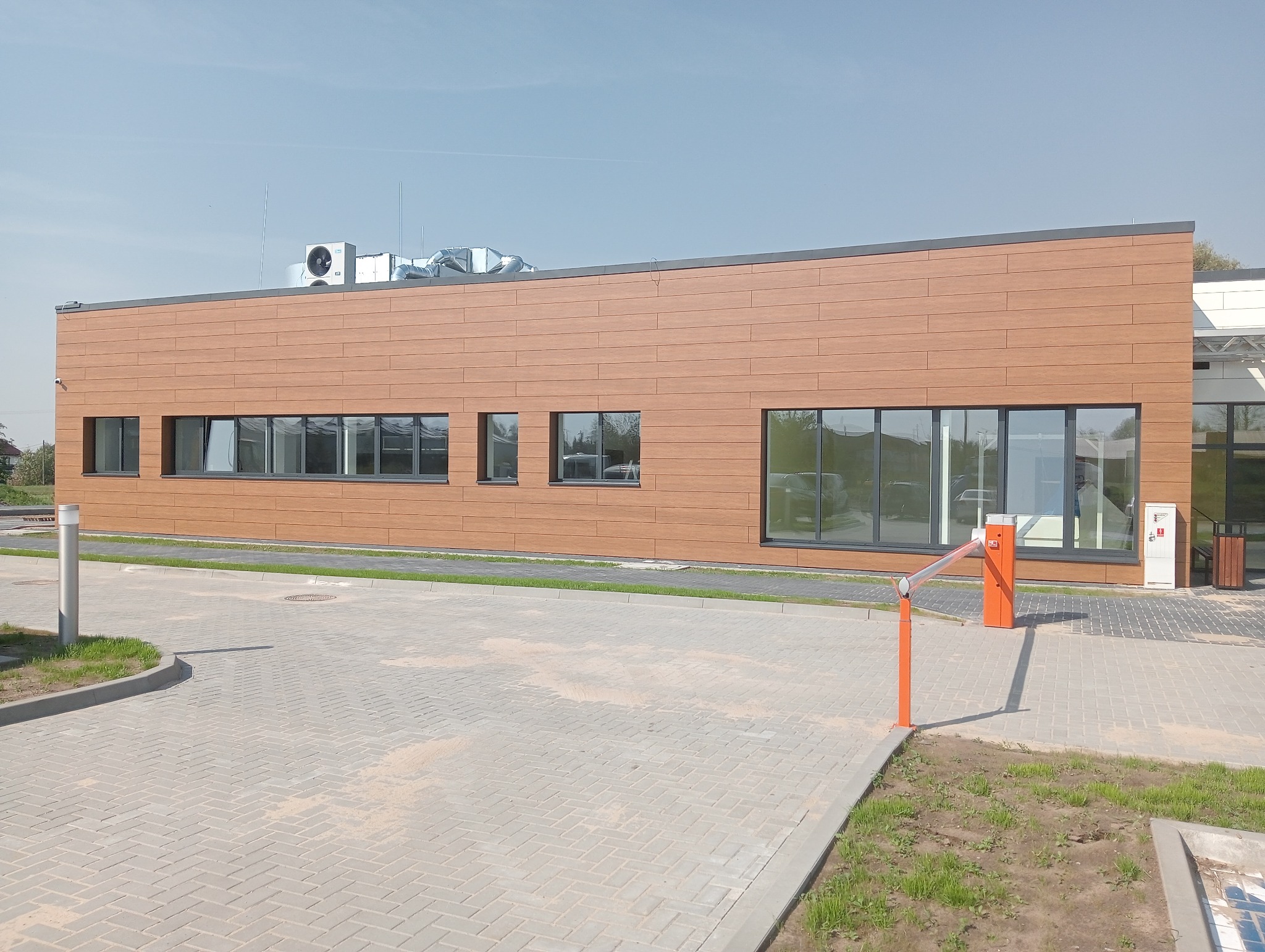 Nowoczesny budynek biurowy z elewacją imitującą drewno, długimi oknami z czarnymi ramami i pomarańczowym szlabanem wjazdowym na pierwszym planie, widok z zewnątrz w słoneczny dzień.