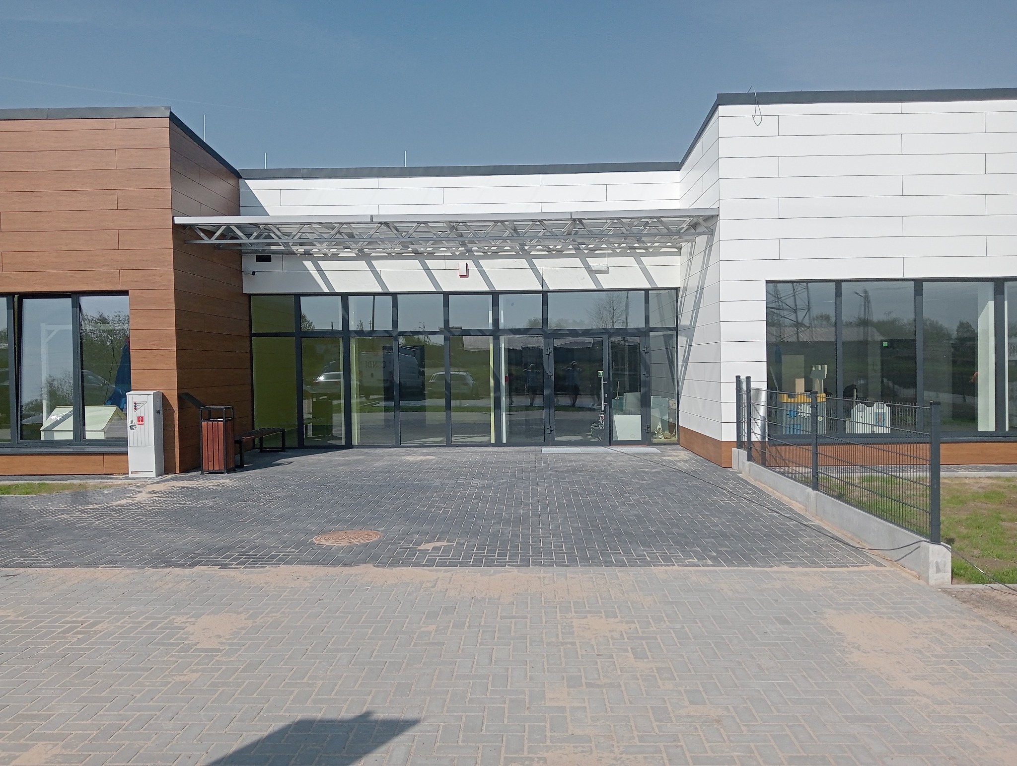 Nowoczesny budynek z fasadą z paneli drewnopodobnych i białych, z dużymi, przeszklonymi drzwiami wejściowymi pod metalową konstrukcją zadaszenia, widok z zewnątrz na brukowany plac.