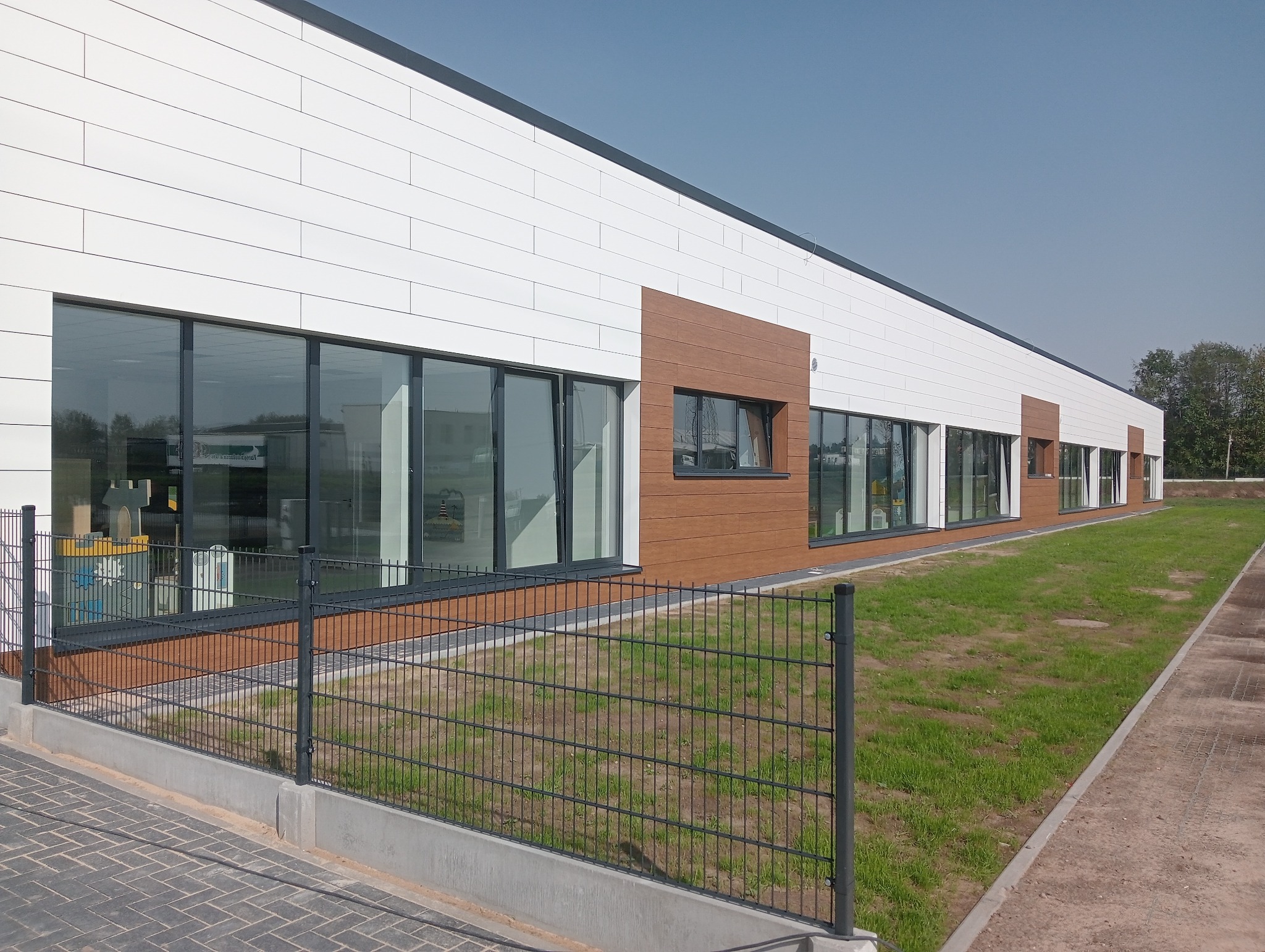 Nowoczesny budynek z białymi panelami elewacyjnymi i brązowymi akcentami, duże przeszklenia z widocznymi zabawkami dziecięcymi wewnątrz, ogrodzenie panelowe z przodu.
