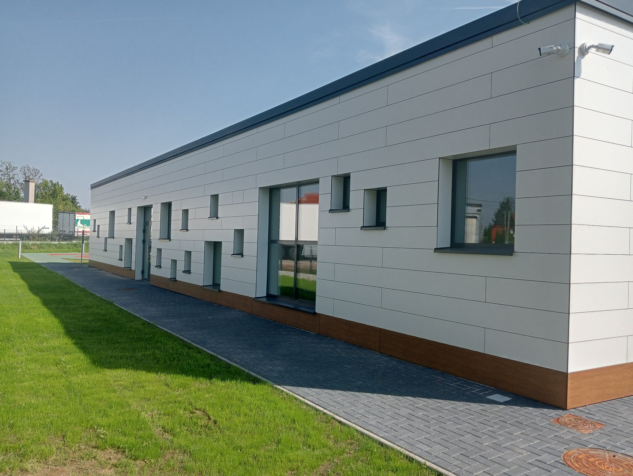Nowoczesny budynek z białymi panelami elewacyjnymi, dużymi przeszkleniami okiennymi i ciemnymi ramami, z widocznymi kamerami monitoringu na elewacji, otoczony trawnikiem i chodnikiem z kostki...