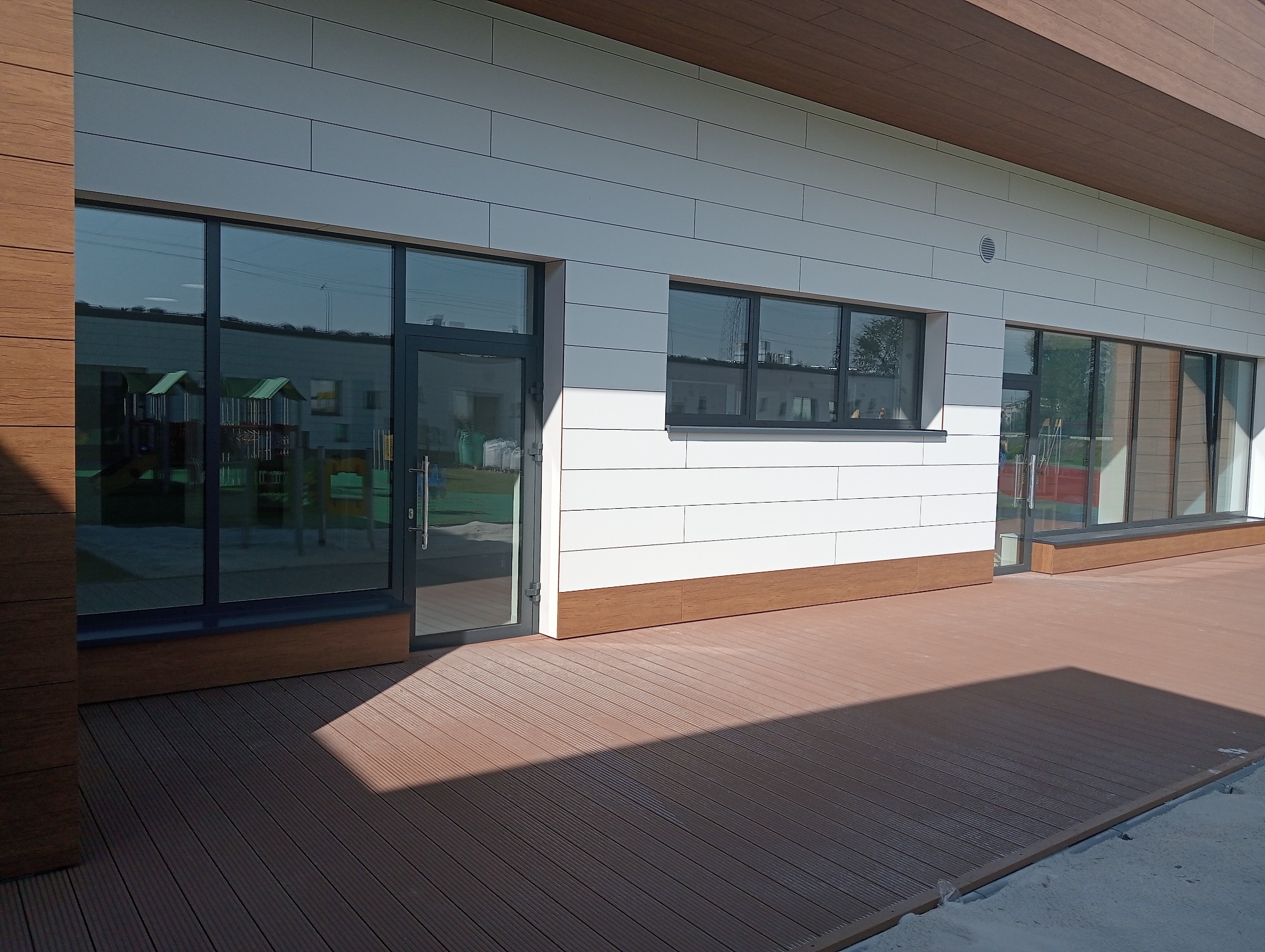 Nowoczesny budynek z białymi panelami elewacyjnymi, grafitową stolarką okienną i drzwiową, oraz tarasem z desek kompozytowych w kolorze brązu. W oknach odbija się plac zabaw.