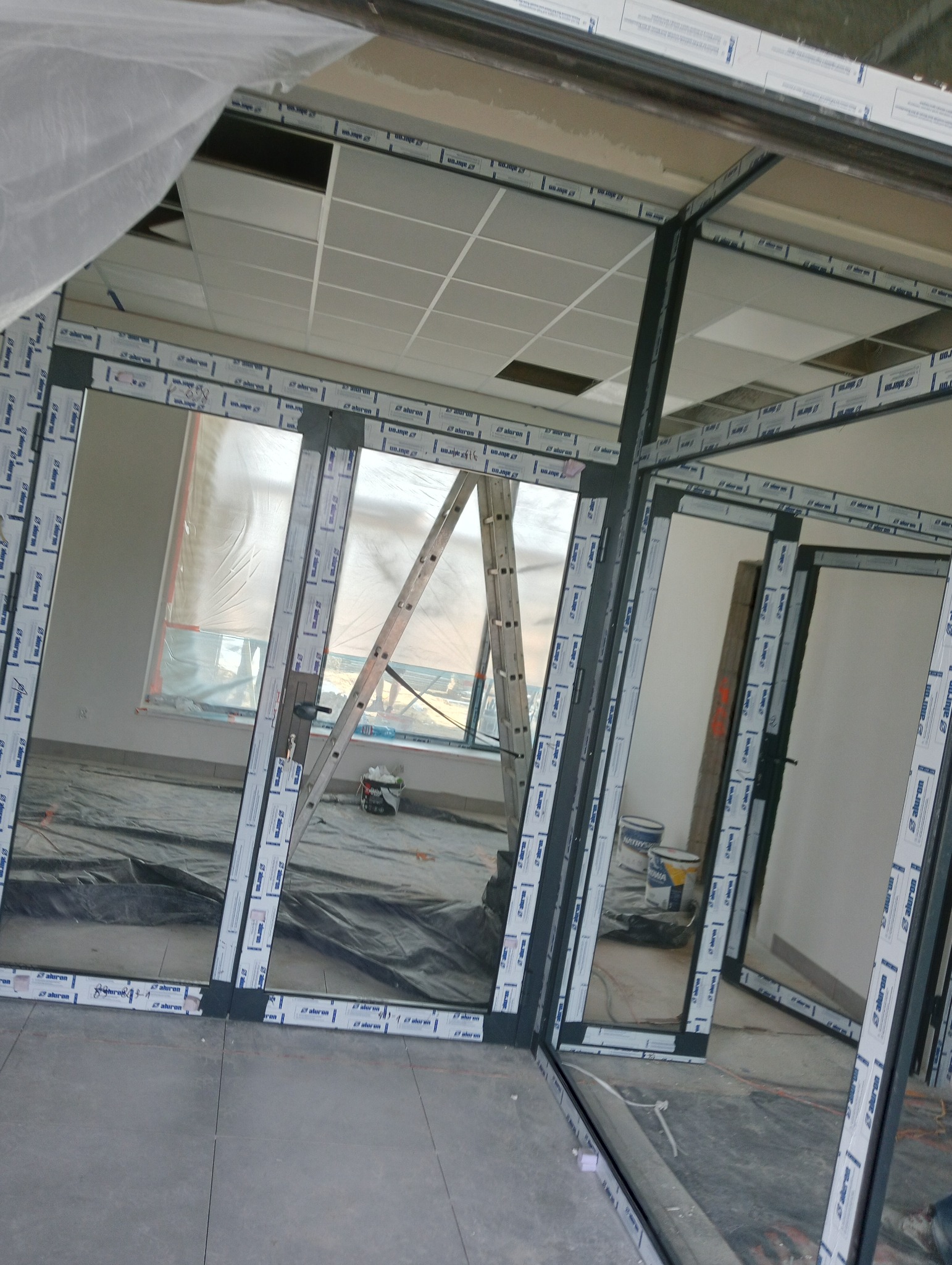 Montaż konstrukcji z profili aluminiowych w surowym wnętrzu, widoczna drabina i zabezpieczenia, ślady prac wykończeniowych na podłodze.