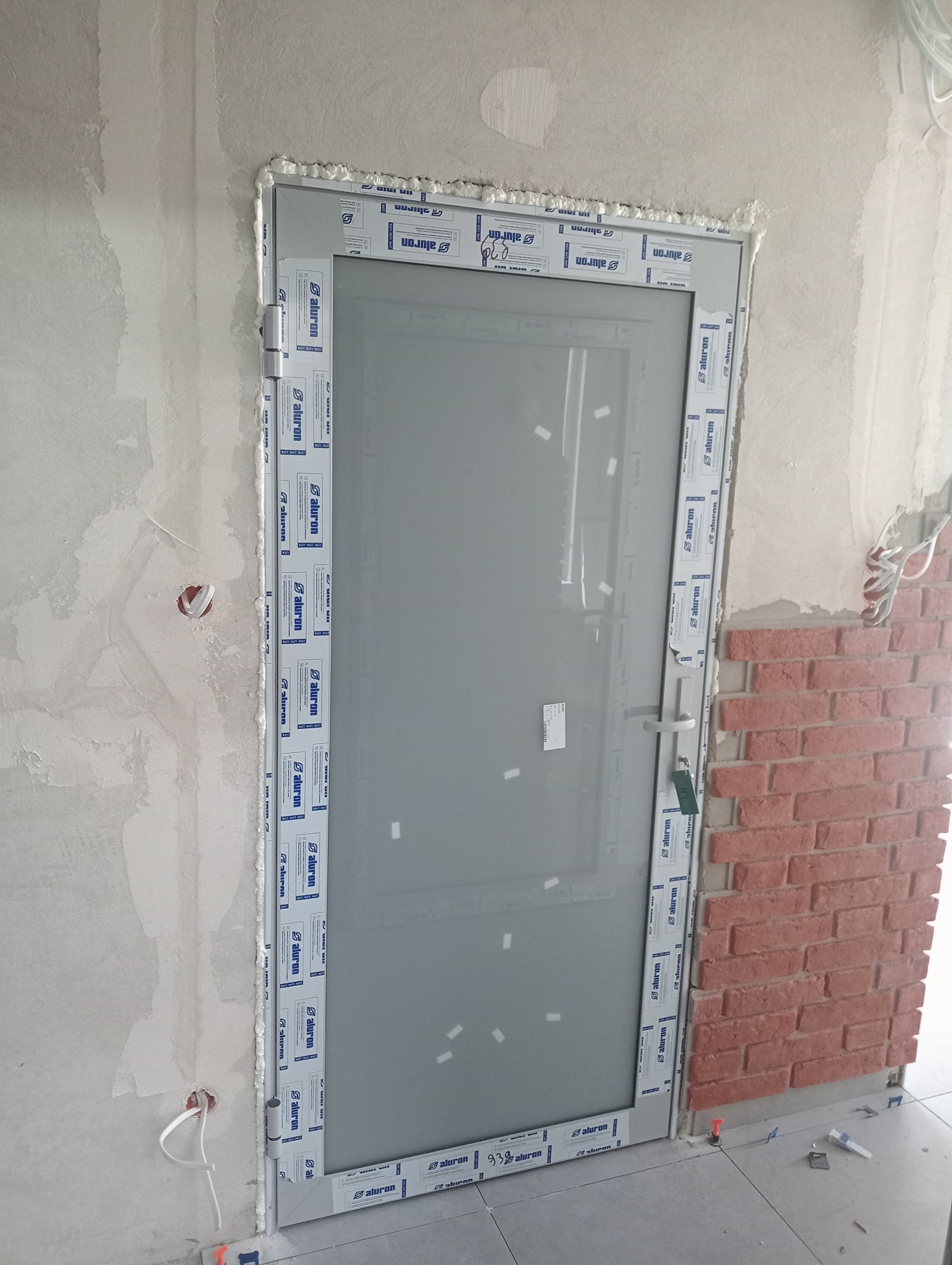 Szara przeszklona konstrukcja osadzona w surowym otworze ściennym, zabezpieczona pianką montażową i folią z logo producenta, z fragmentem ściany wyłożonej cegłą dekoracyjną w tle.