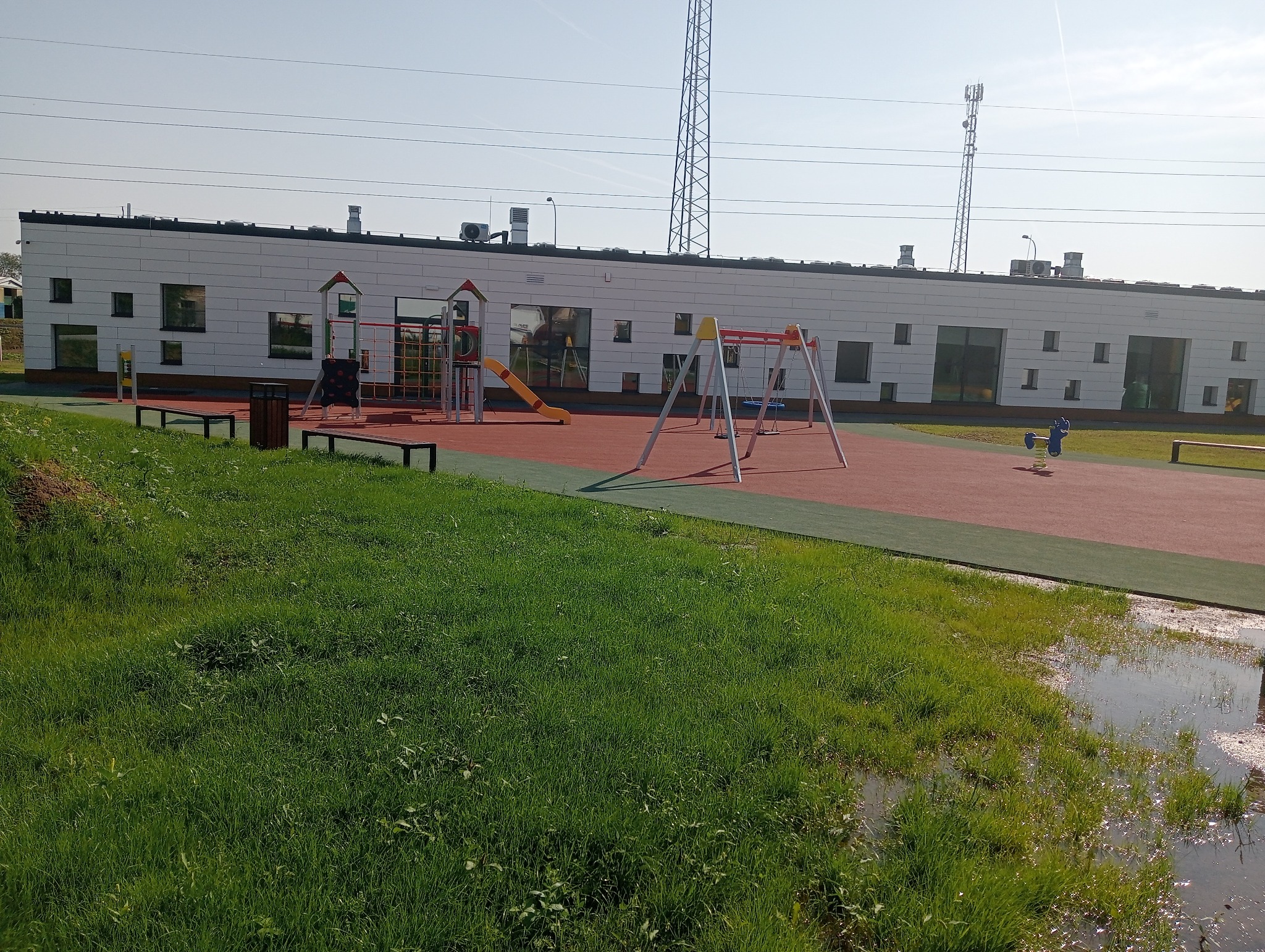 Nowoczesny plac zabaw z huśtawkami, zjeżdżalnią i sprężynowym konikiem na czerwonej nawierzchni, otoczony zielonym trawnikiem i budynkiem z licznymi oknami o różnych rozmiarach.