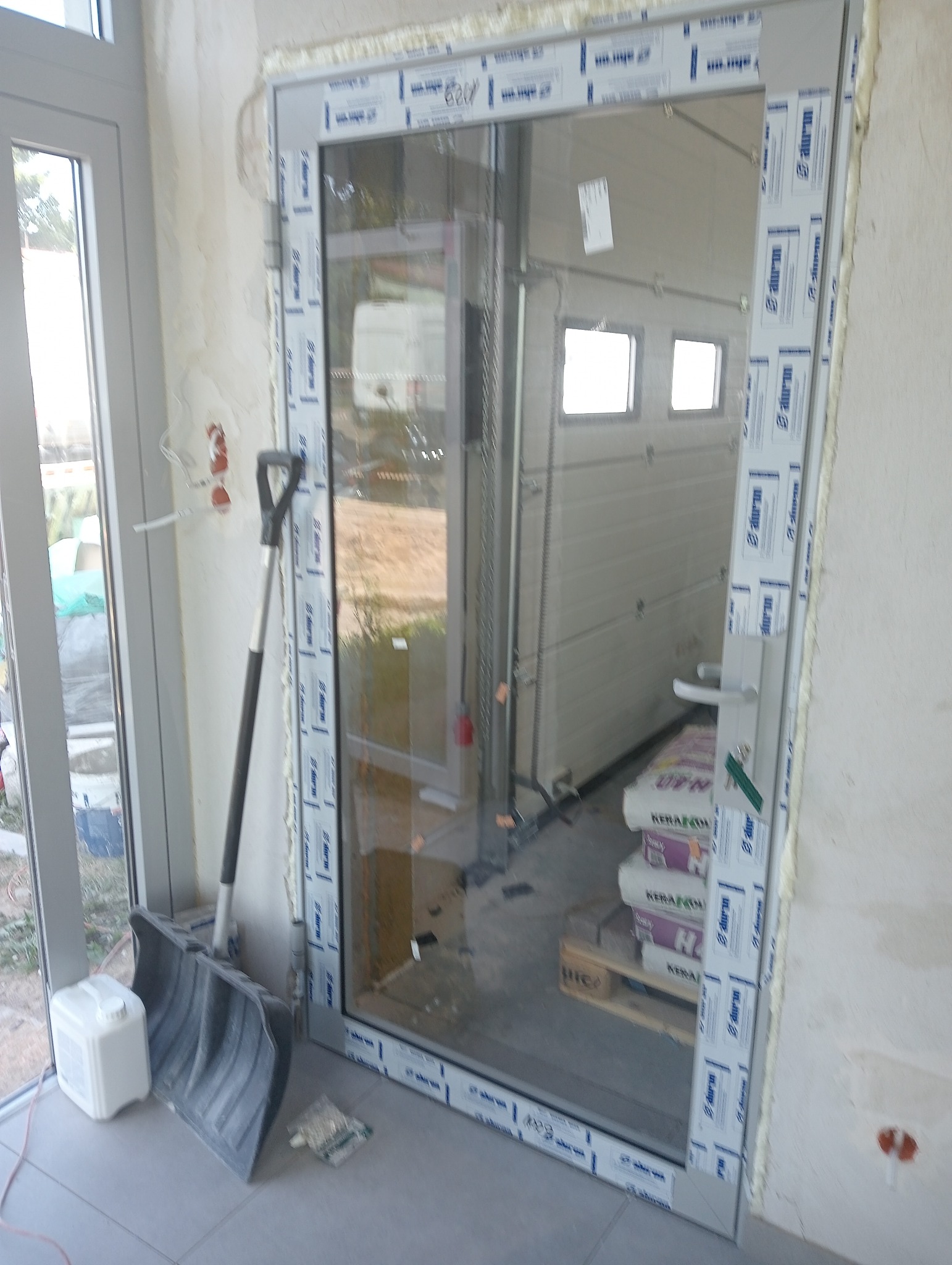 Montaż okna z widoczną częścią bramy garażowej w tle, obok stoi szufelka i kanister, ściany w stanie surowym.