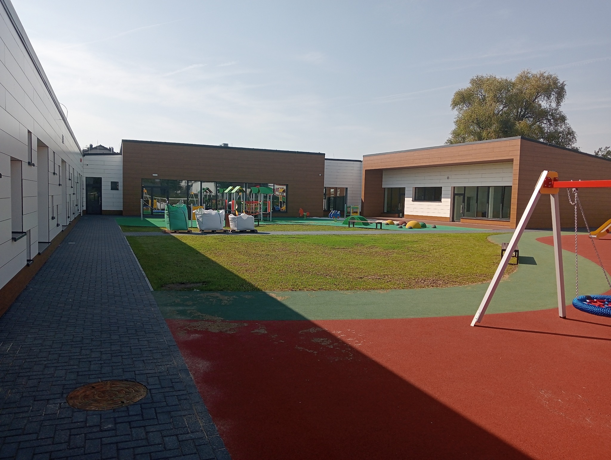 Nowoczesny budynek przedszkola z placem zabaw na zewnątrz, widoczne okna z ciemnymi ramami, kolorowa nawierzchnia placu zabaw i elementy wyposażenia