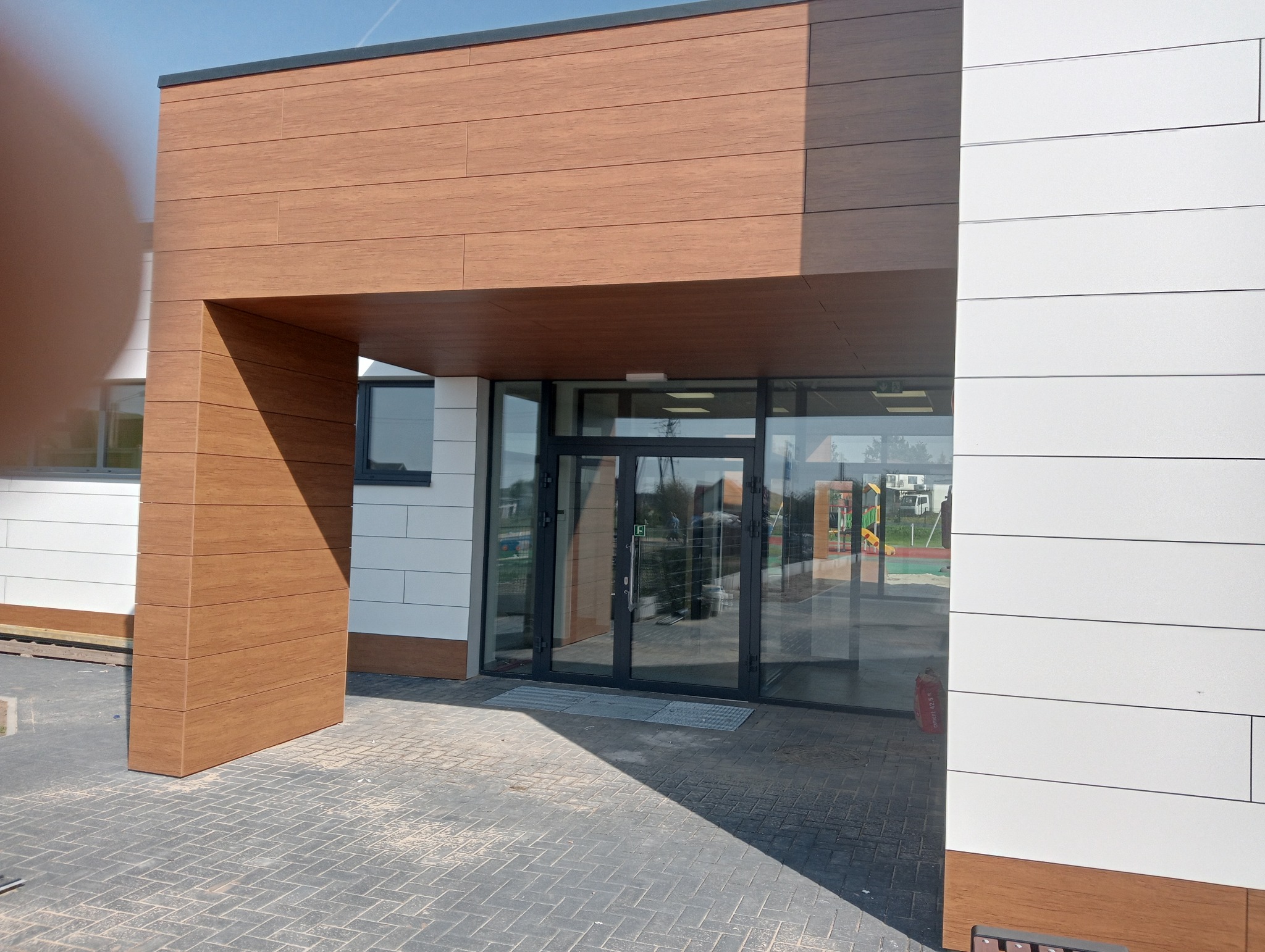 Nowoczesne, przeszklone drzwi wejściowe do budynku w Choroszczy, z okładziną elewacyjną imitującą drewno i białą, poziomą okładziną, widoczne odbicie placu zabaw w szybach.