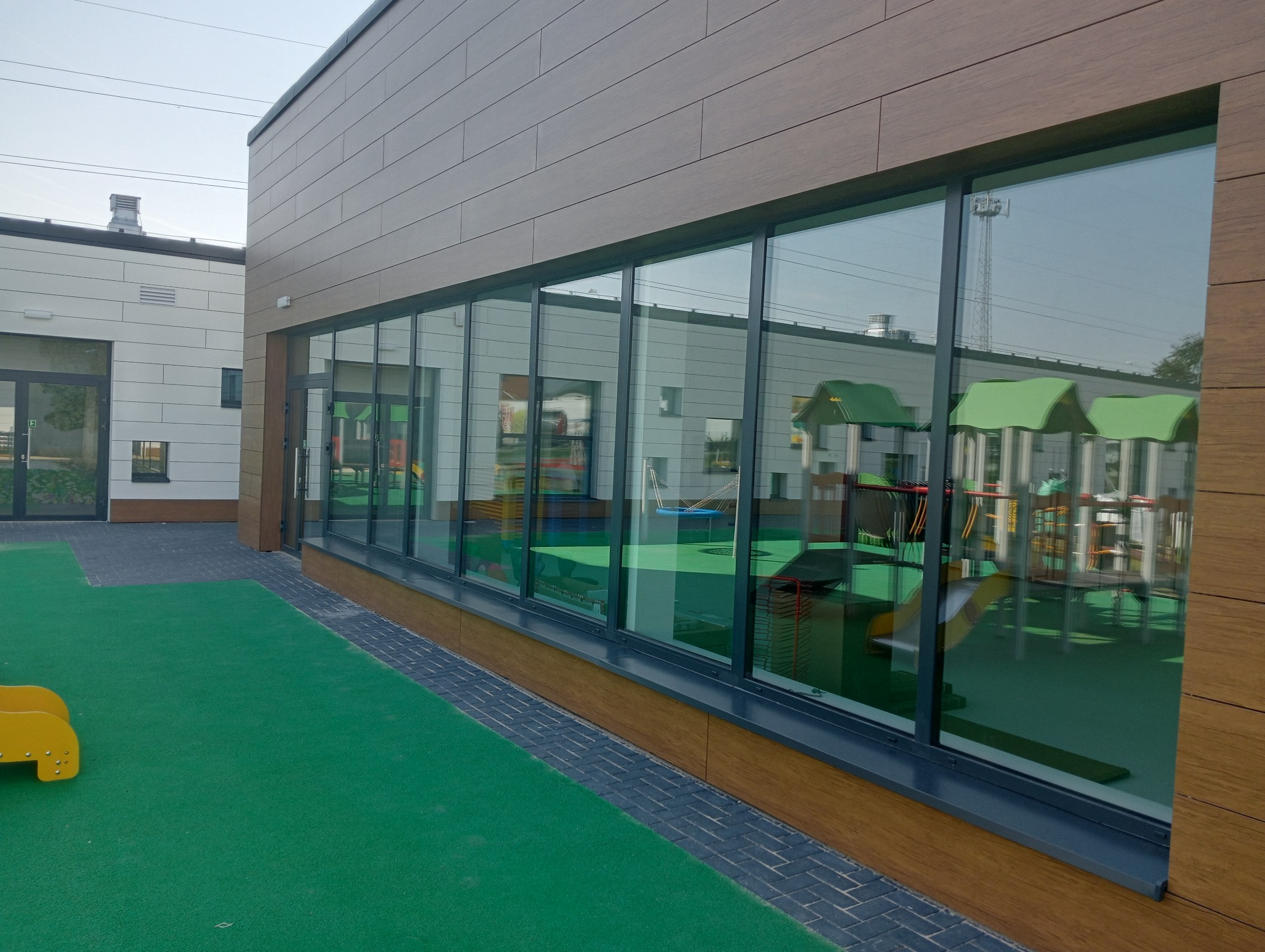 Duże, panoramiczne przeszklenia w budynku przedszkola z drewnopodobną elewacją, odbijające elementy placu zabaw.