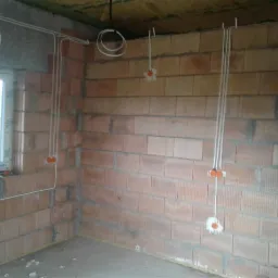 Wewnętrzna instalacja elektryczna w surowym budynku: widoczne przewody w peszlach zamocowane do ściany z czerwonej cegły, puszki instalacyjne z pomarańczowymi dekielkami.