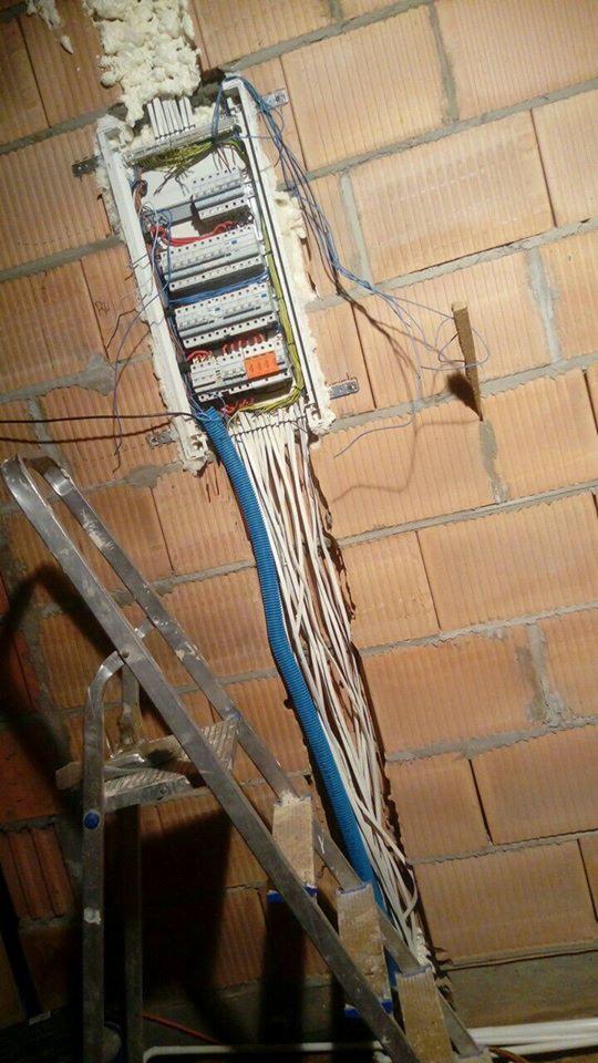 Rozdzielnica elektryczna w trakcie montażu w surowej ścianie z cegły, widoczne kable w niebieskiej rurce karbowanej i drabina aluminiowa.