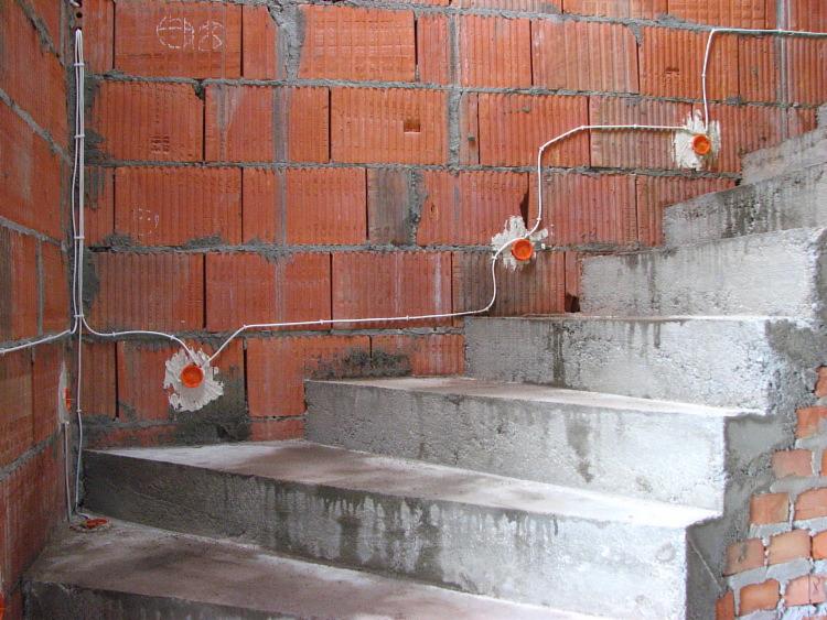 Surowy stan: Instalacja elektryczna w trakcie montażu na ceglanej ścianie klatki schodowej, widoczne przewody i puszki instalacyjne.