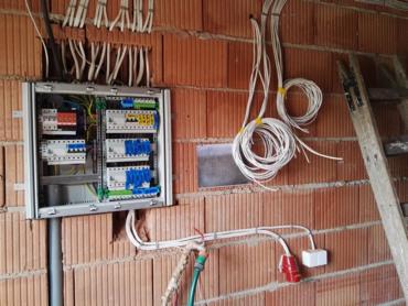 Rozdzielnica elektryczna zamontowana na surowej ścianie z cegły dziurawki, widoczne przewody, drabina oparta o ścianę, instalacja w trakcie wykonywania.