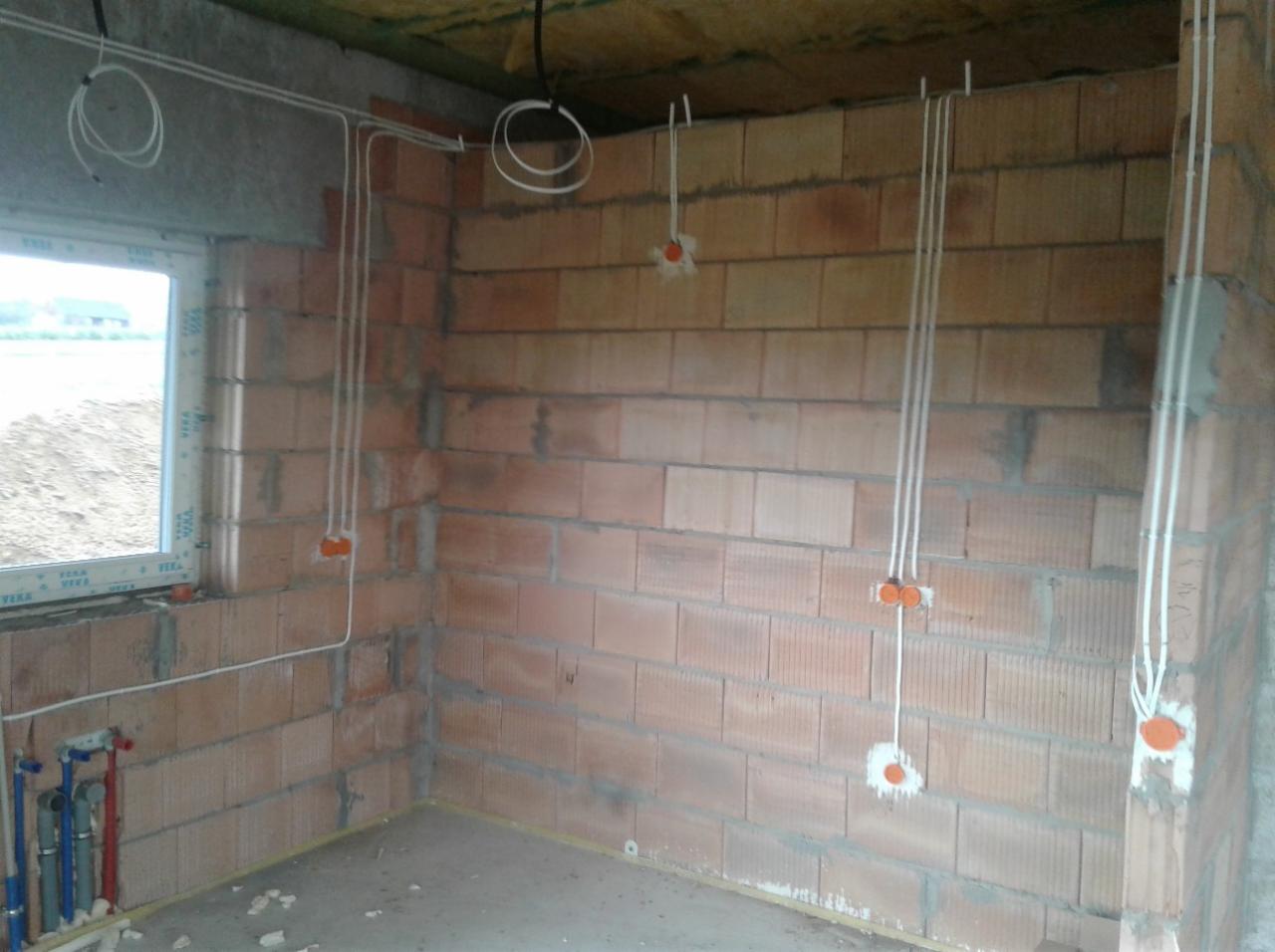 Wewnętrzna instalacja elektryczna w surowym budynku: widoczne przewody w peszlach zamocowane do ściany z czerwonej cegły, puszki instalacyjne z pomarańczowymi dekielkami.