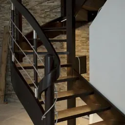 Kręte schody drewniane z metalowymi relingami w domu w Zalewie, oświetlone od dołu, widoczne na tle kamiennej ściany.