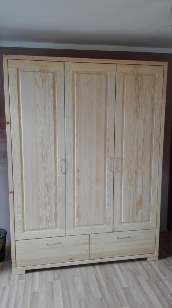 Trzydrzwiowa, jasna, sosnowa szafa z dwoma szufladami na dole, z metalowymi uchwytami, na tle ściany w dwóch odcieniach brązu i szarej podłogi.