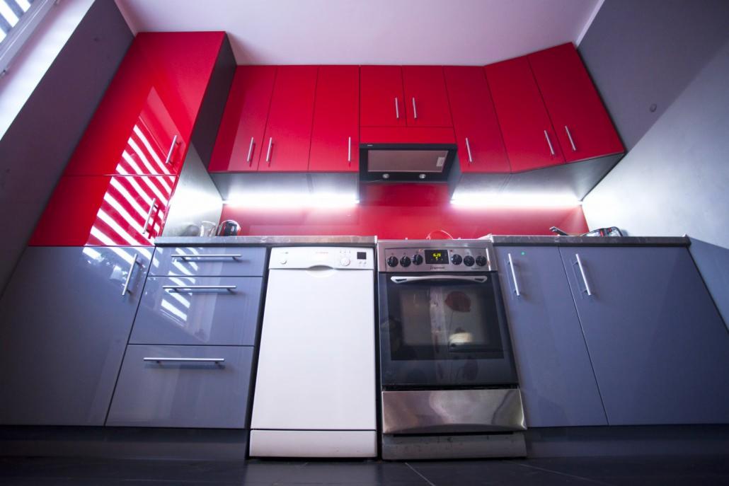 Nowoczesna kuchnia z czerwonymi szafkami górnymi i szarymi dolnymi, oświetlenie LED pod szafkami, zabudowa pod skosem dachu.