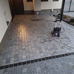 Abra - Szara kostka granitowa ułożona na podjeździe, z czarnym pasem oddzielającym dwa rodzaje kostki. W tle budynek z oknami i myjka ciśnieniowa.