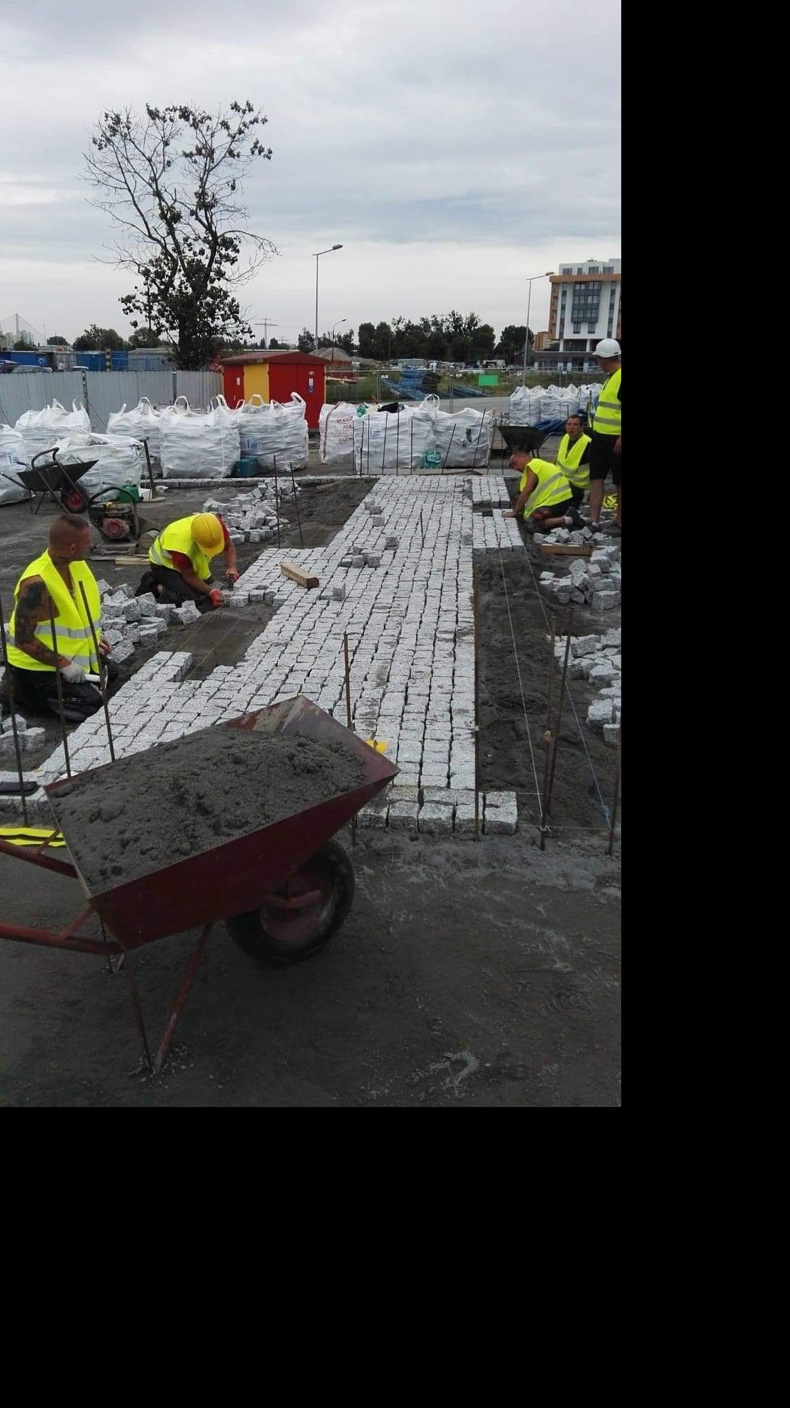 Układanie szarej kostki granitowej na podsypce; w tle wózki z materiałami budowlanymi i pracownicy w żółtych kamizelkach.