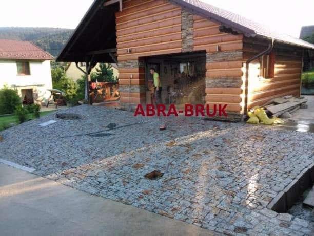 Ułożona szara kostka brukowa przed drewnianym budynkiem z elementami kamiennymi, widoczna osoba w tle i częściowo ułożony obszar z drobnym żwirem.