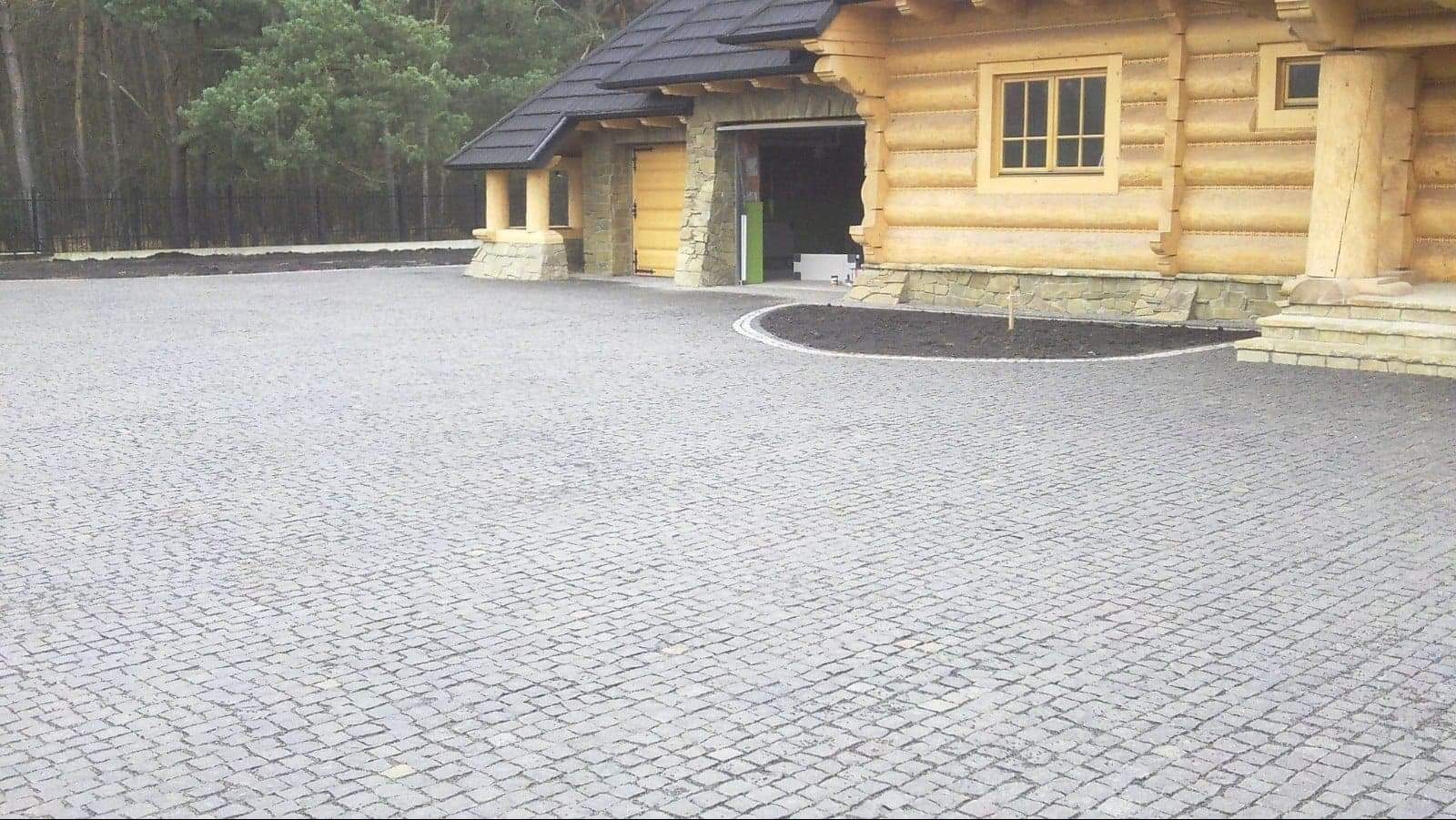 Ułożona szara kostka granitowa na podjeździe przed drewnianym domem z garażem, widoczny fragment obrzeża z kostki.
