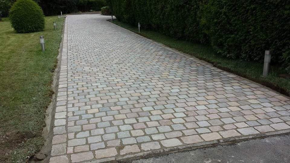 Długa, wybrukowana kostką granitową droga dojazdowa, obramowana trawnikiem i żywopłotem, oświetlona niskimi lampami.