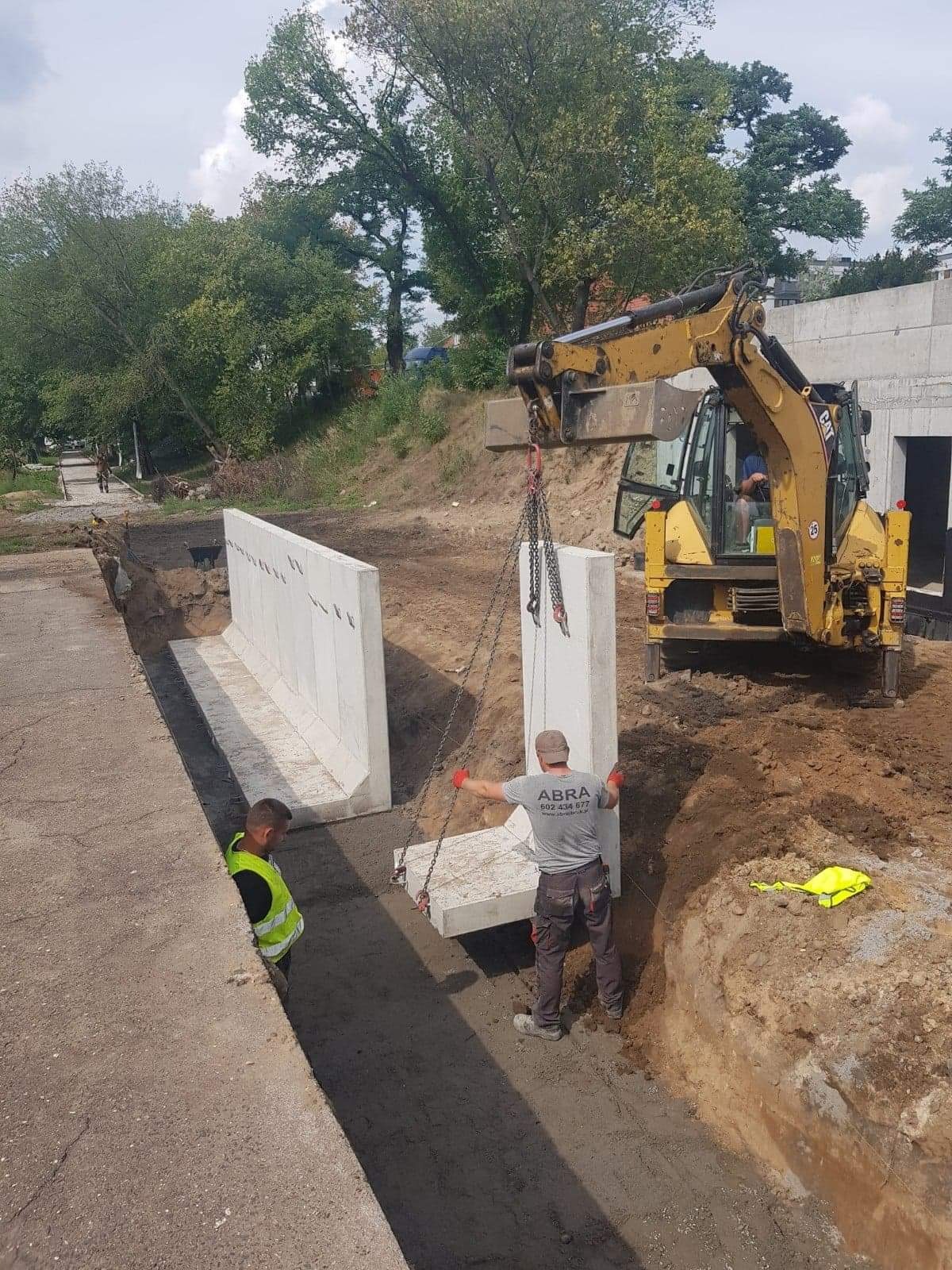 Żółta koparko-ładowarka CAT umieszcza betonowy element w wykopie, dwaj pracownicy w kamizelkach odblaskowych nadzorują proces, widoczny fragment chodnika i zieleń w tle.