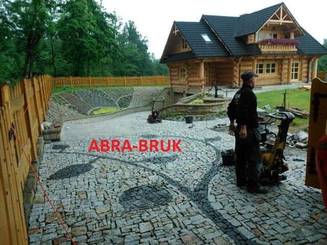 Ukończona nawierzchnia z kostki brukowej ułożonej we wzór liści, z pracownikiem i ubijarką w tle, obok drewnianego domu i ogrodzenia.