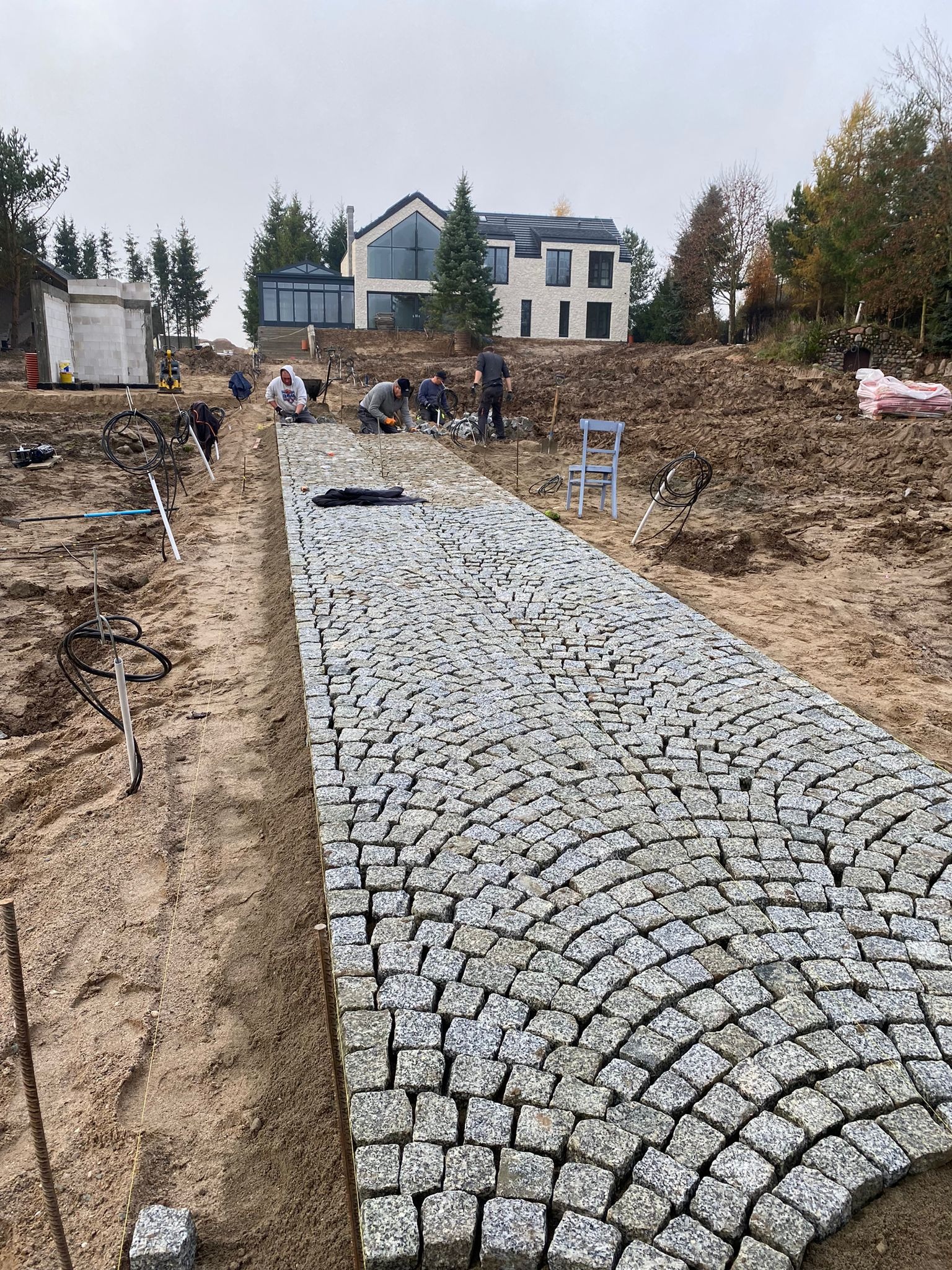 Ekipa budowlana układa ścieżkę z kostki granitowej w łukowaty wzór na posesji z nowoczesnym domem w tle, widoczne narzędzia i materiały budowlane.