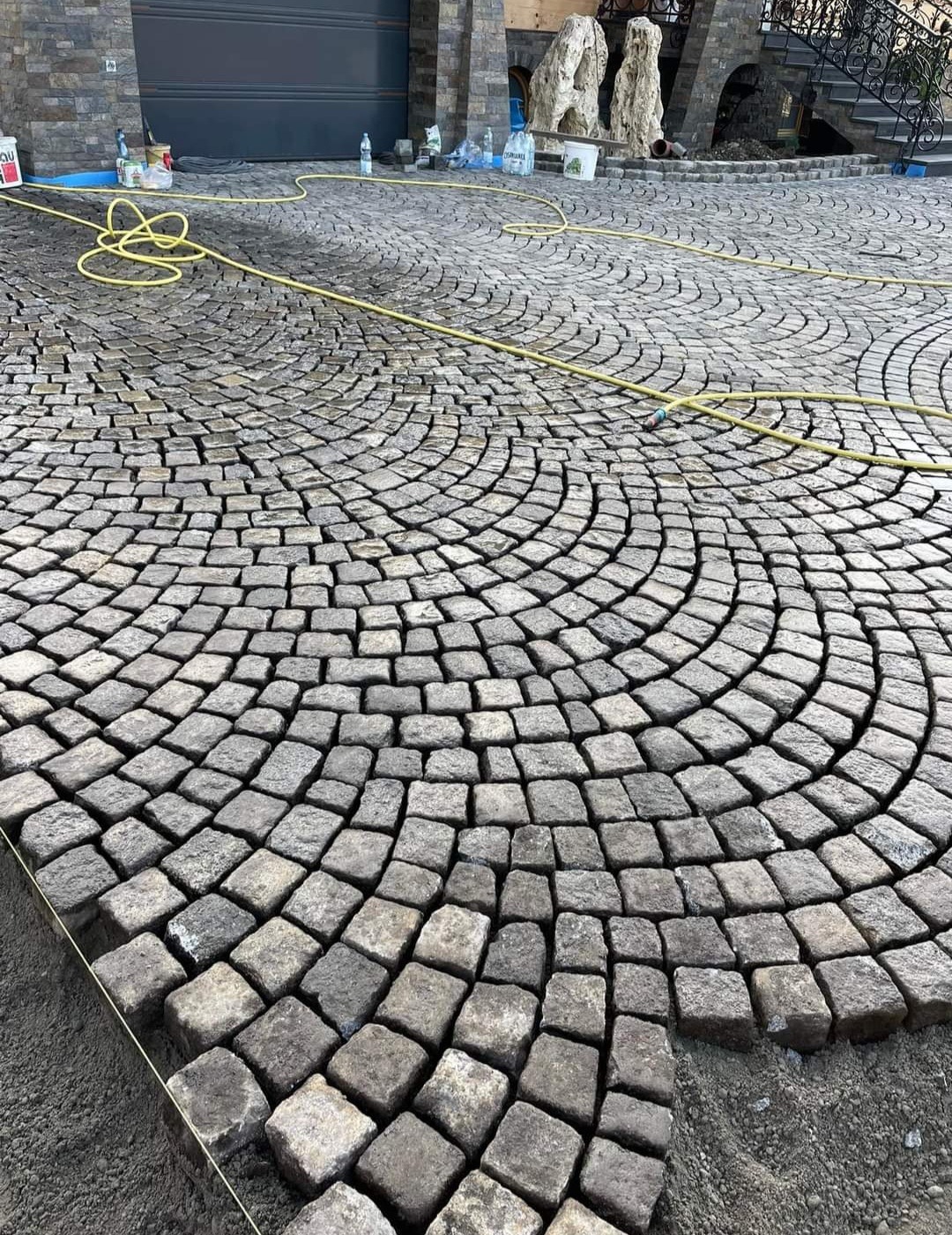 Półokrągły wzór z szarej kostki brukowej na podjeździe, widoczne narzędzia i materiały budowlane w tle.