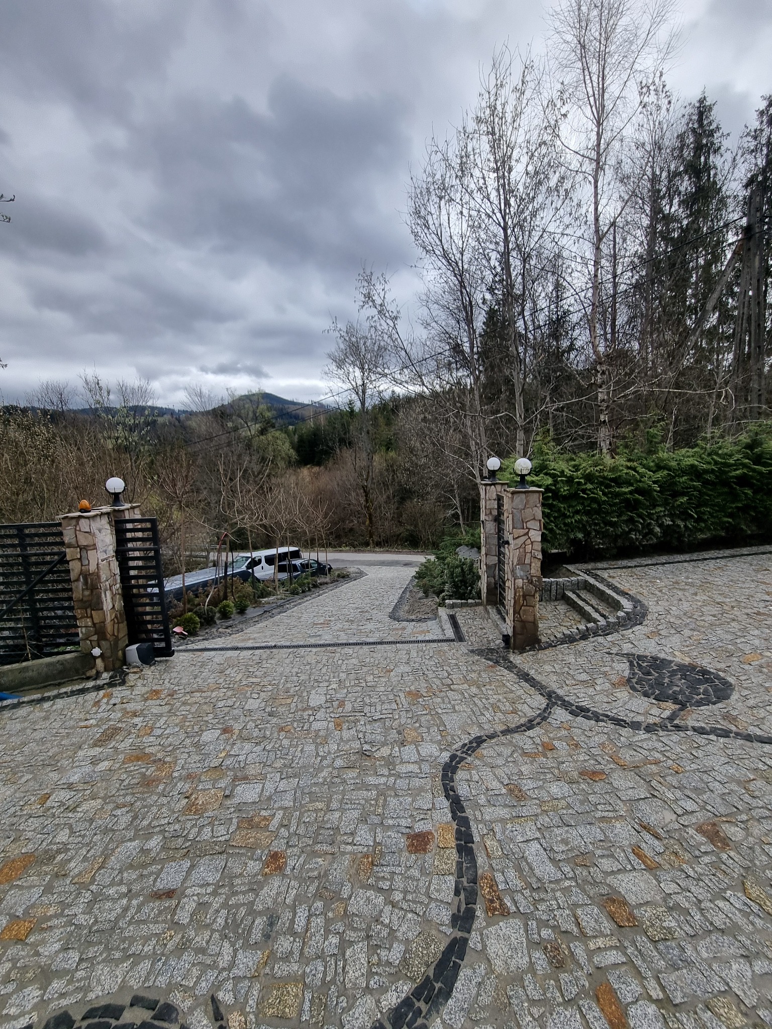 Podjazd wyłożony szarą kostką z ciemnymi wzorami, brama wjazdowa z kamiennymi słupkami zwieńczonymi lampami, w tle zalesione wzgórza pod pochmurnym niebem.