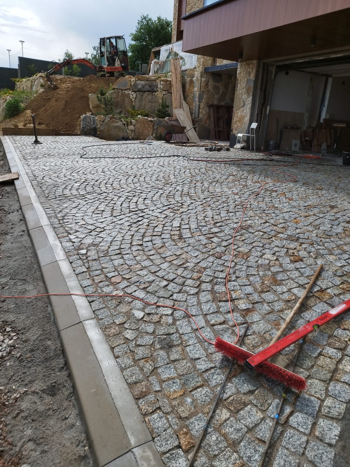 Ułożona, łukowata nawierzchnia z kostki granitowej w odcieniach szarości, ograniczona betonowym obrzeżem. W tle widoczny budynek z garażem, wykop ziemny i koparka.