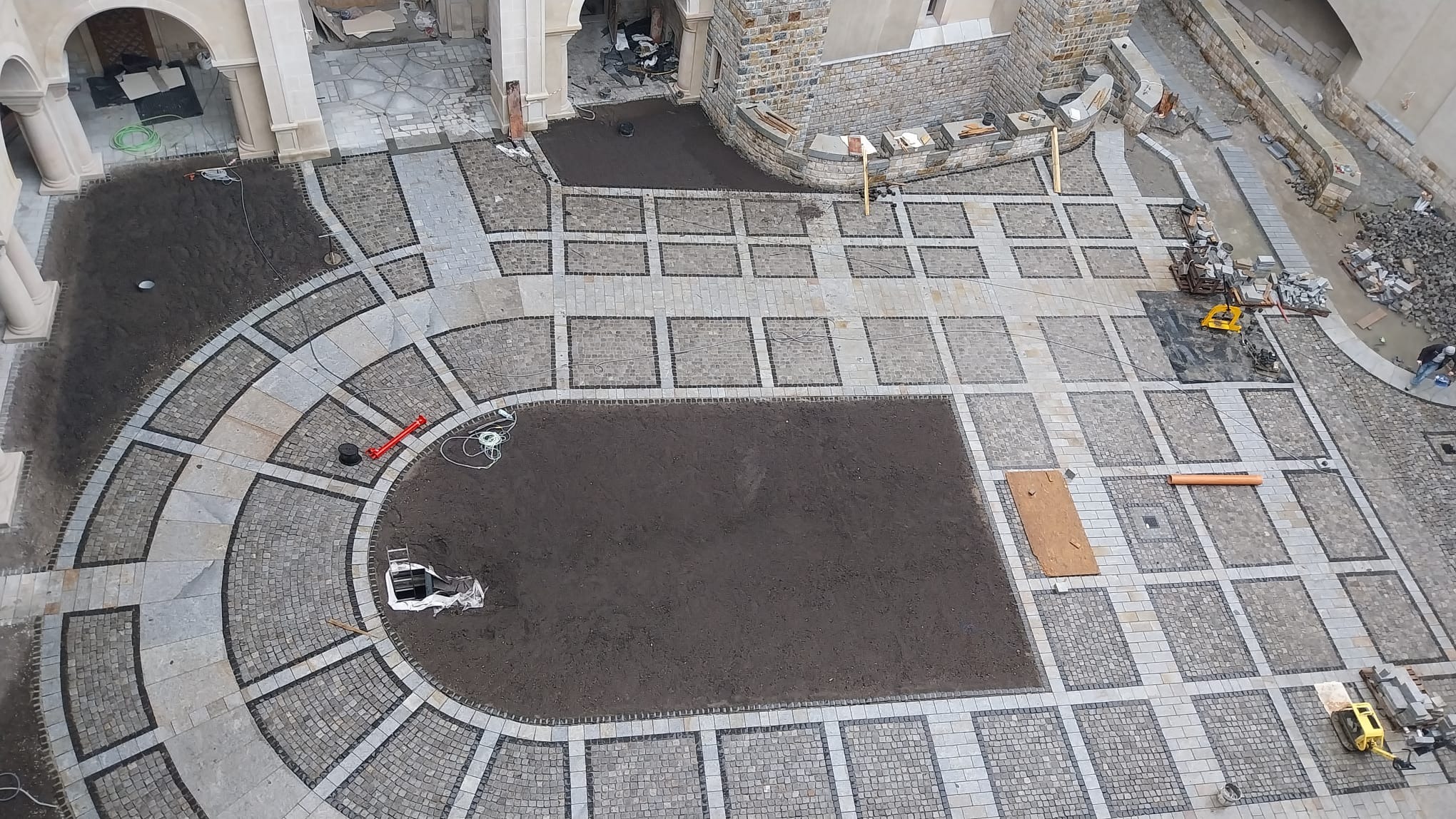 Widok z góry na plac z częściowo ułożoną kostką w geometryczne wzory, z fragmentami ziemi przygotowanymi pod dalsze prace, widoczne narzędzia i materiały budowlane.