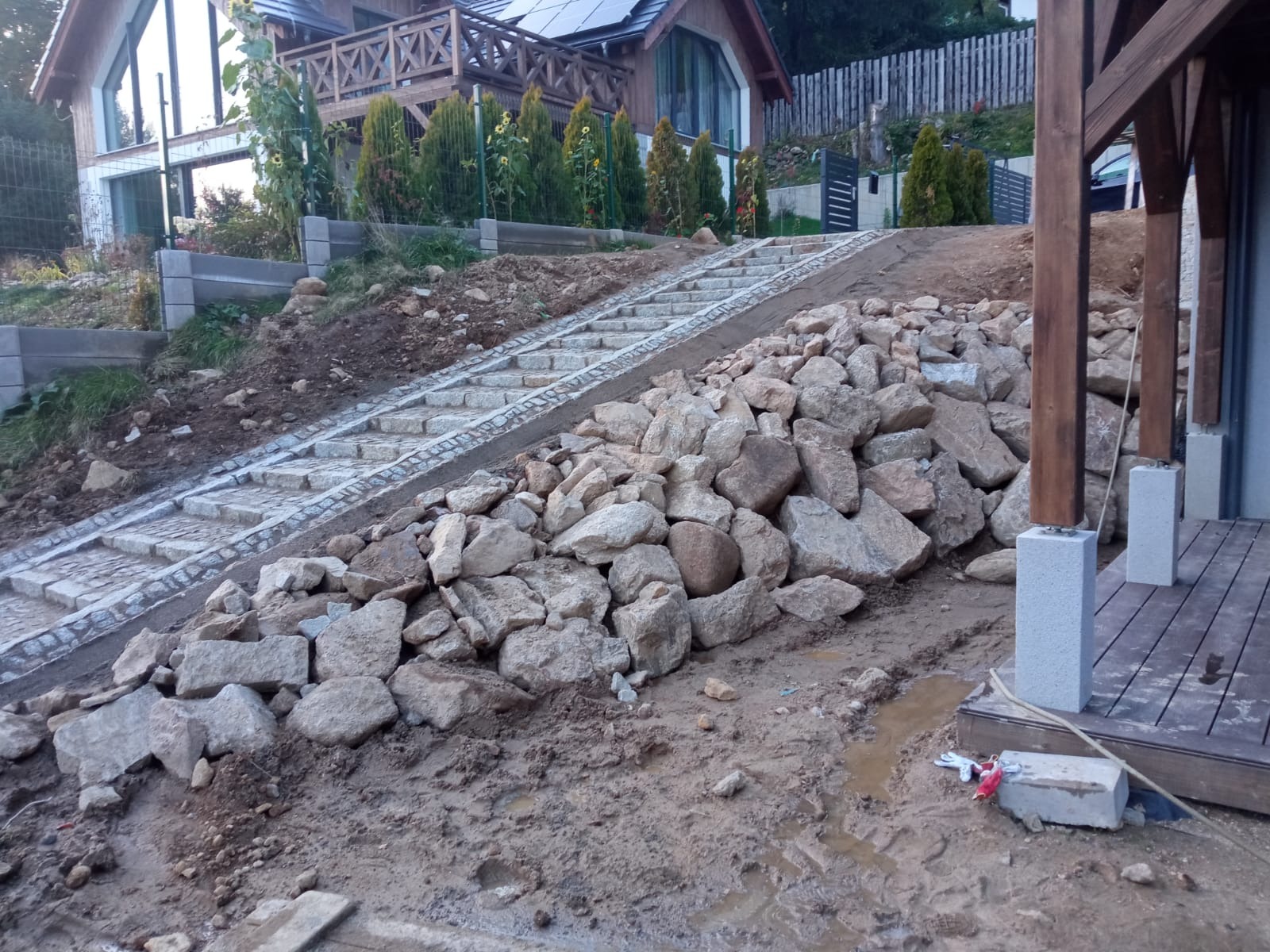 Kamienne schody prowadzące do domu z drewnianym balkonem, częściowo obsypane dużymi kamieniami, widoczne ślady prac ziemnych.