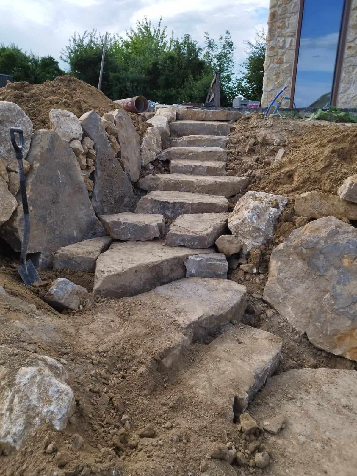 Kamienne schody ogrodowe w trakcie budowy, z widoczną łopatą opartą o duży głaz, ziemia i roślinność w tle.