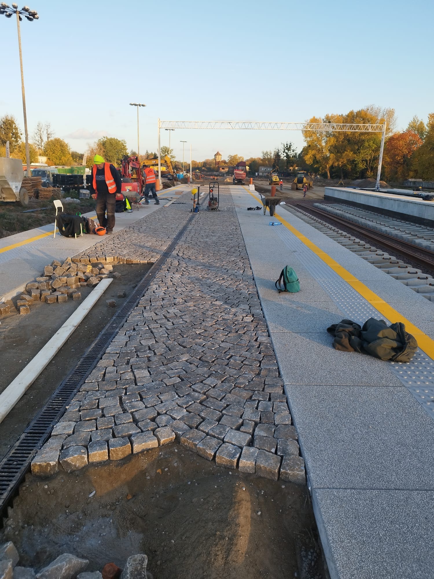 Fragment budowy peronu kolejowego z częściowo ułożoną kostką granitową, widoczni pracownicy w odblaskowych kamizelkach, maszyny budowlane w tle.