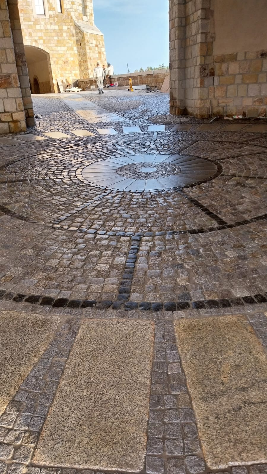 Dziedziniec z okrągłym wzorem ułożonym z kostki granitowej w odcieniach szarości i czerni, z fragmentami jasnych płyt kamiennych, widok z perspektywy niskiego kąta w kierunku łuku kamiennej budowli...