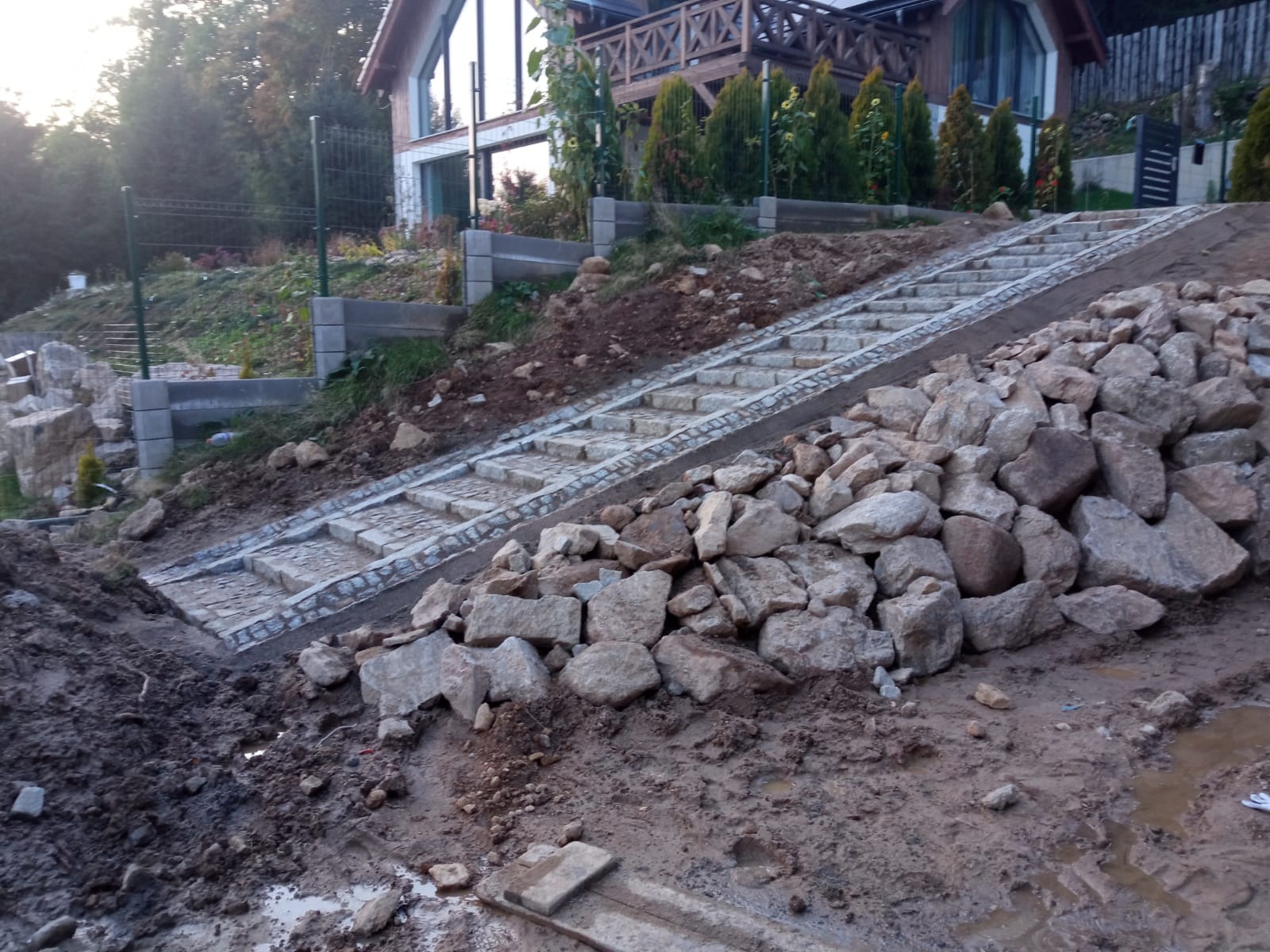 Kamienne schody prowadzące do domu na wzgórzu, częściowo ukończone, z hałdą kamieni u podnóża i błotnistym terenem.