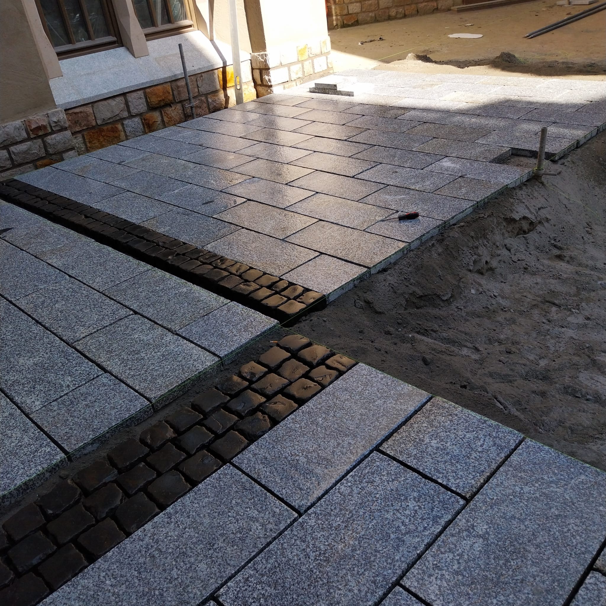 Fragment układanej nawierzchni z prostokątnych, szarych płyt granitowych, oddzielonych pasem z ciemnej kostki brukowej, widoczny fragment przygotowanego podłoża.