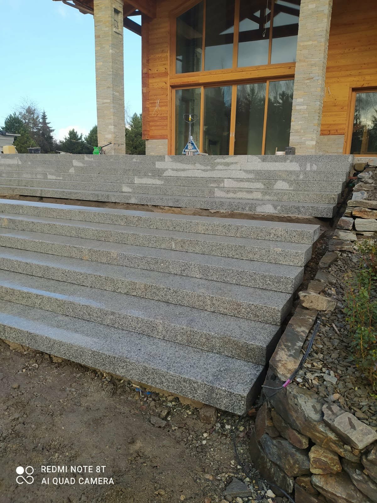 Granitowe schody wejściowe do domu z drewnianą elewacją, częściowo wykończone, z widocznymi elementami montażowymi i kamiennym obrzeżem.
