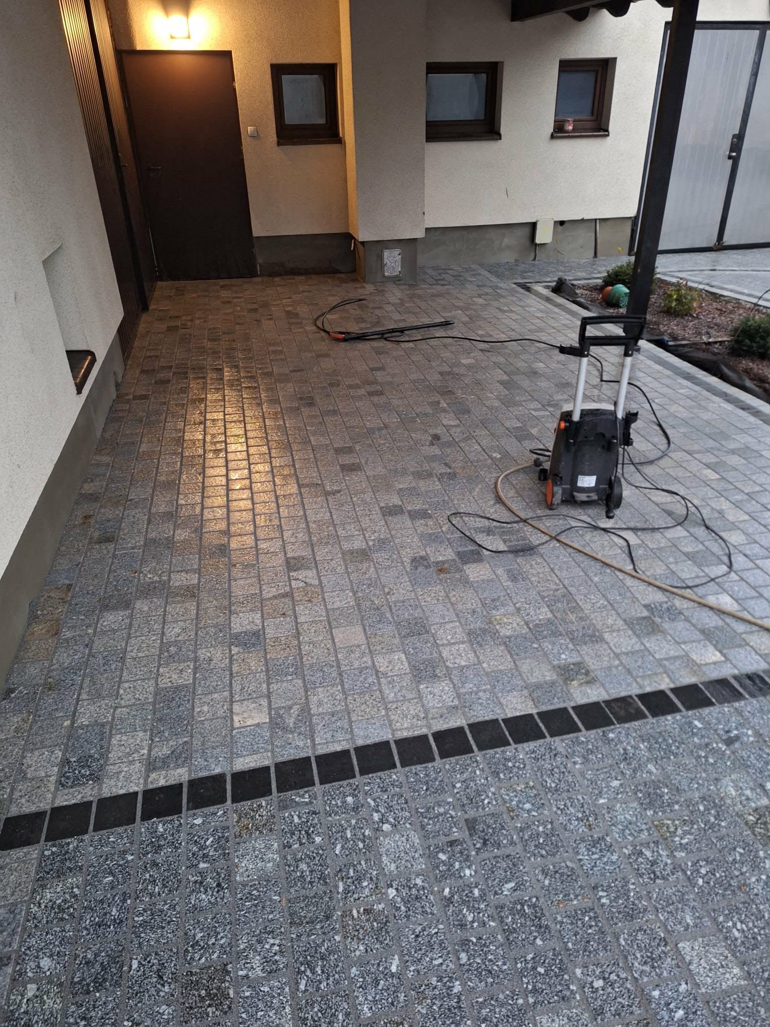 Szara kostka granitowa ułożona na podjeździe, z czarnym pasem oddzielającym dwa rodzaje kostki. W tle budynek z oknami i myjka ciśnieniowa.