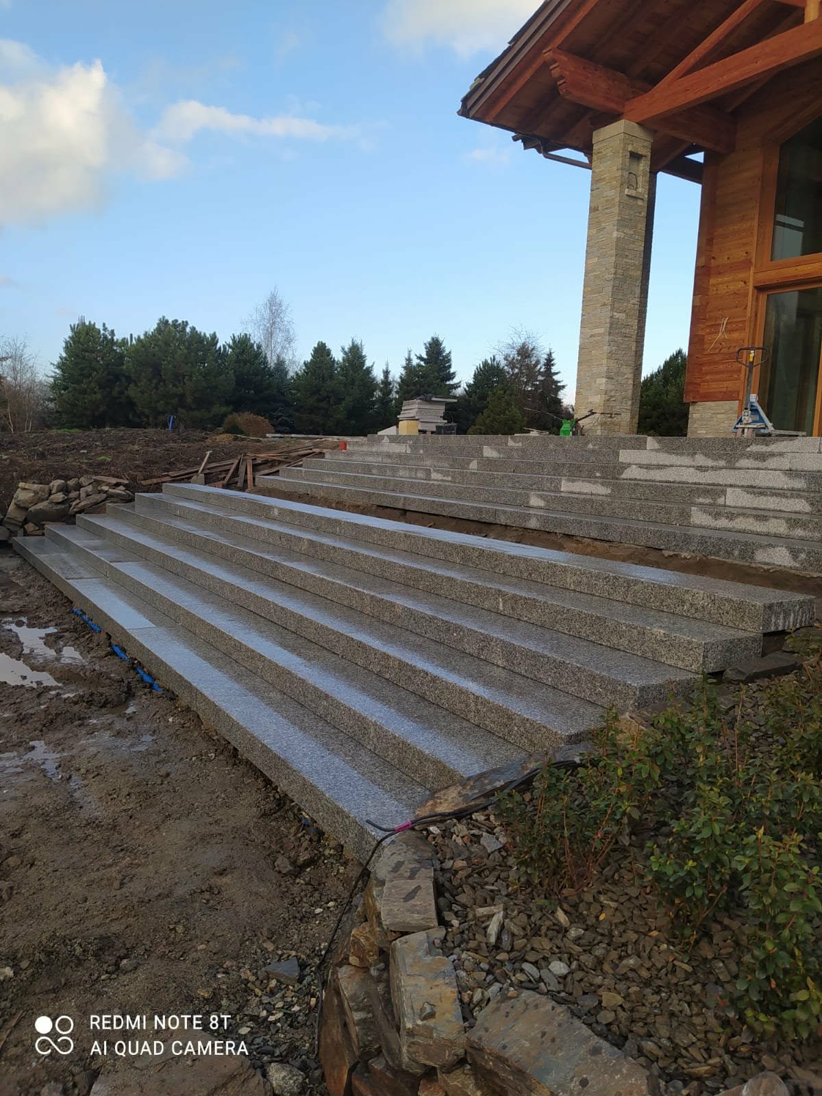 Granitowe schody wejściowe do domu w trakcie budowy, widoczne elementy konstrukcyjne i otoczenie placu budowy. Ujęcie z dołu, podkreślające perspektywę.